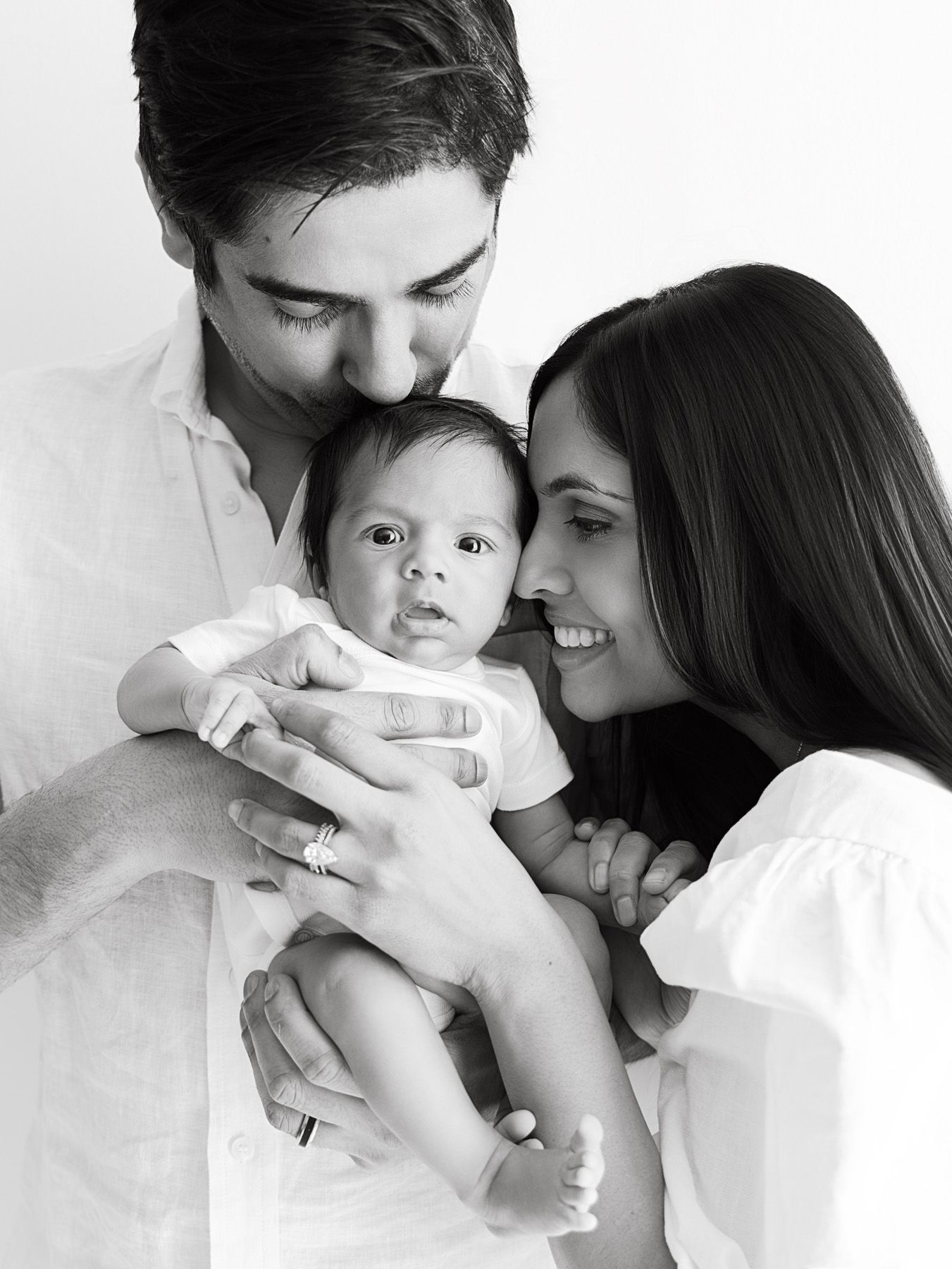 Black and white photo of parents lovingly holding their newborn baby, smiling warmly in a bright studio setting.