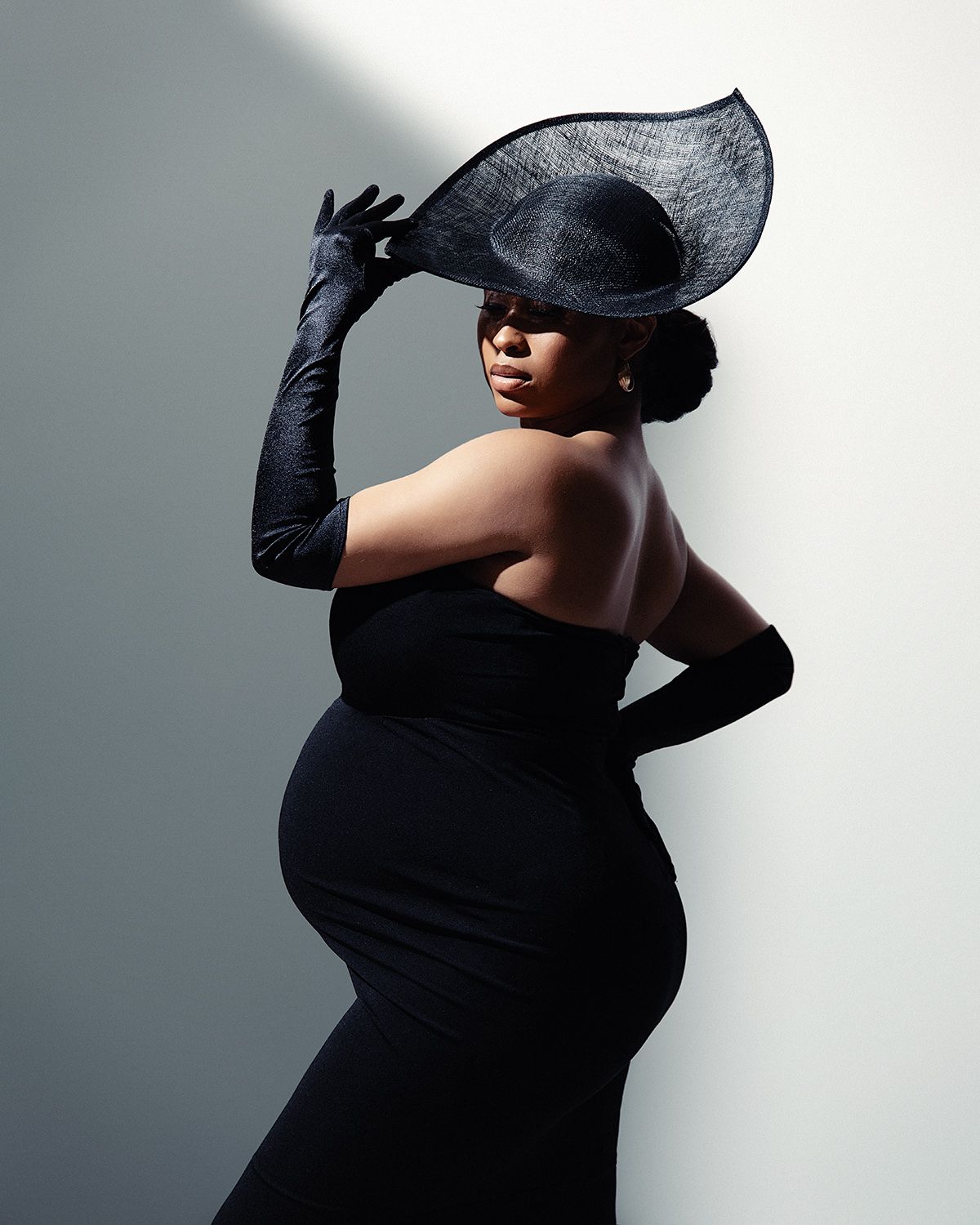 Elegant pregnant woman in black strapless gown, wide-brim hat, and long gloves posing gracefully against a white wall.