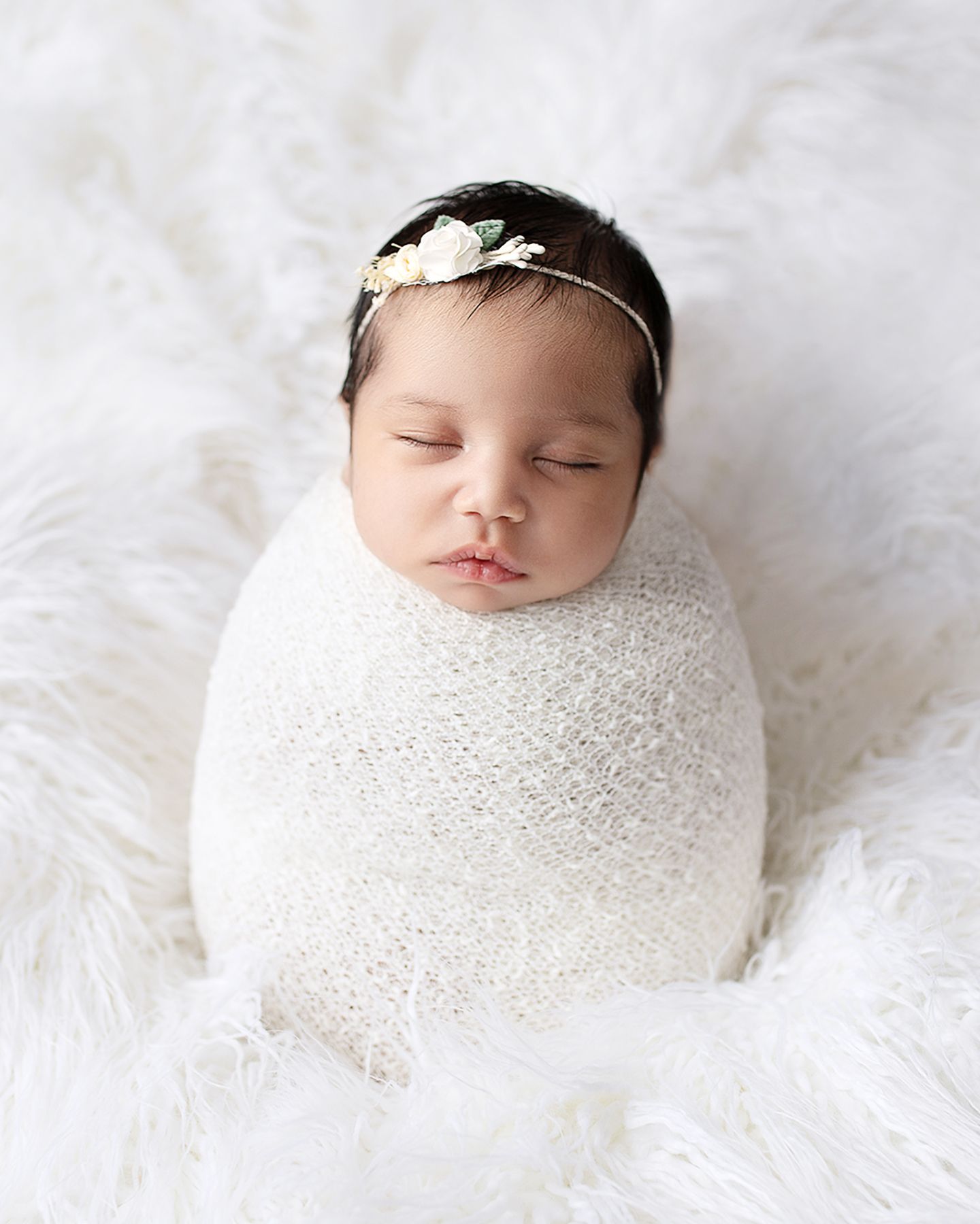 Sleeping newborn baby swaddled in white wrap with floral headband on fluffy white fur background.