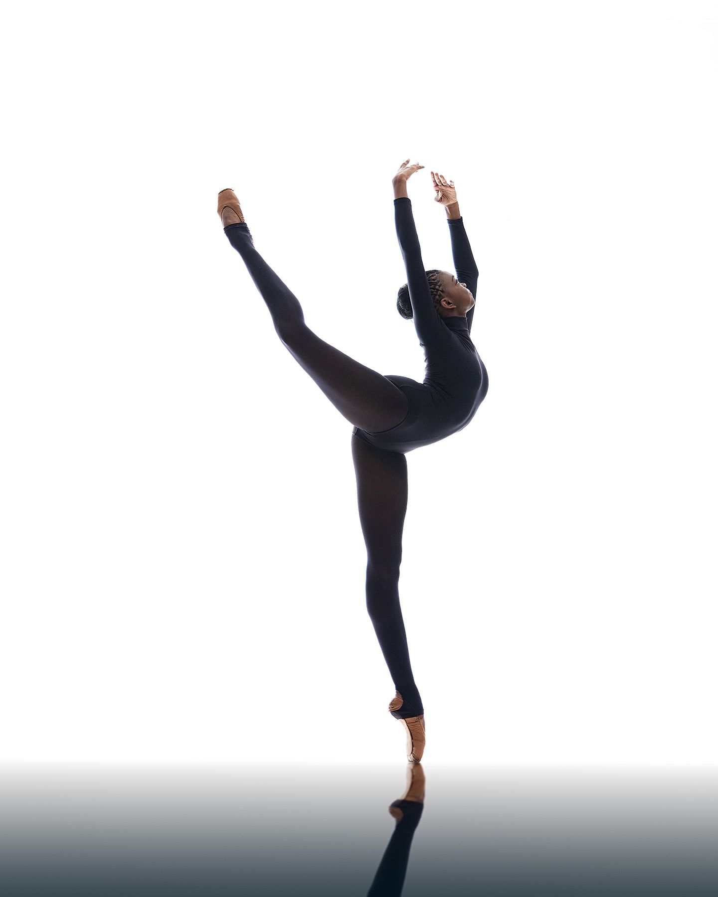 A dancer performs a graceful high leg extension while wearing black dance attire against a neutral background.
