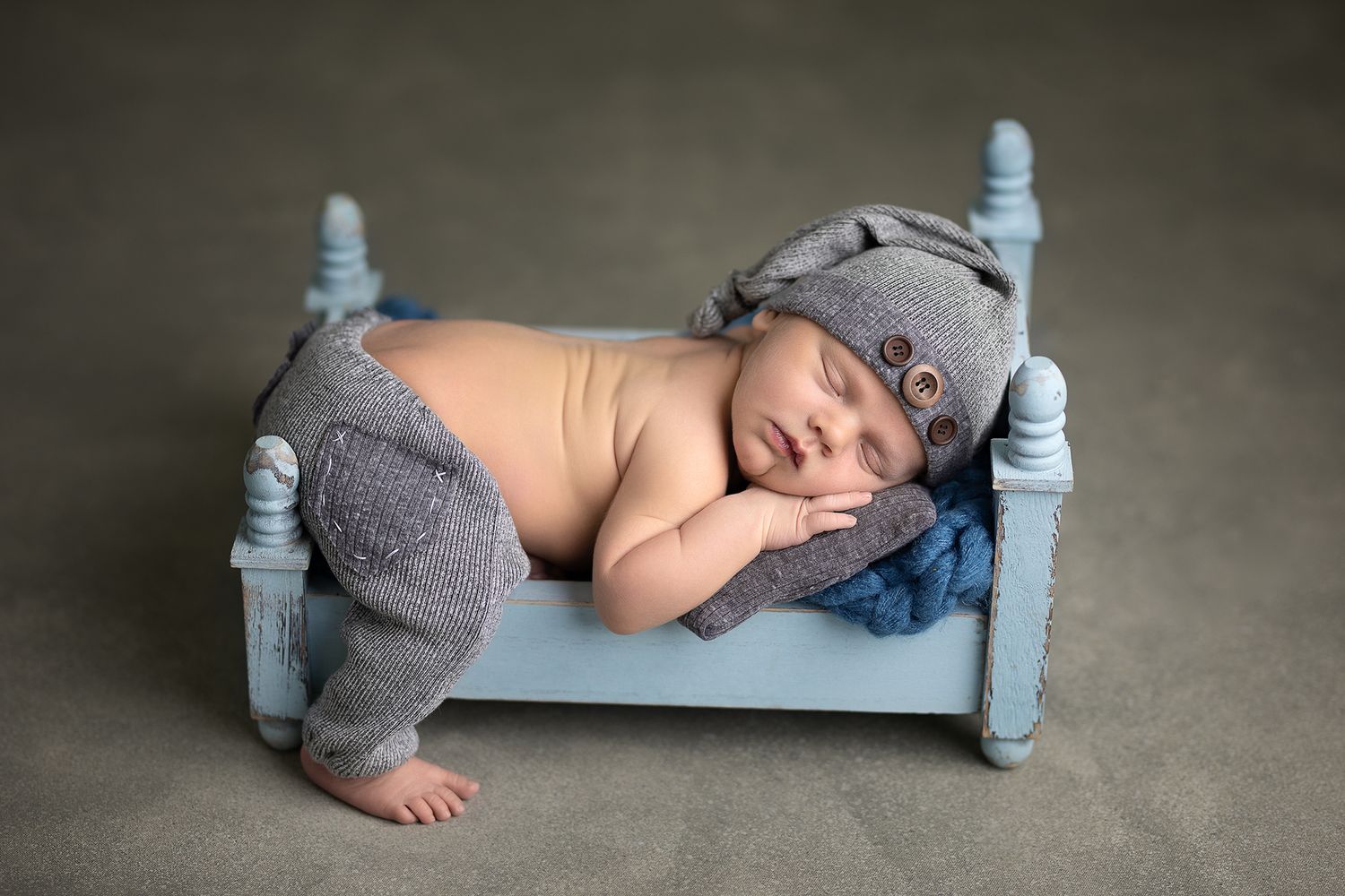 Newborn baby sleeping peacefully on a tiny blue bed wearing gray knit pants and hat in a professional photo.
