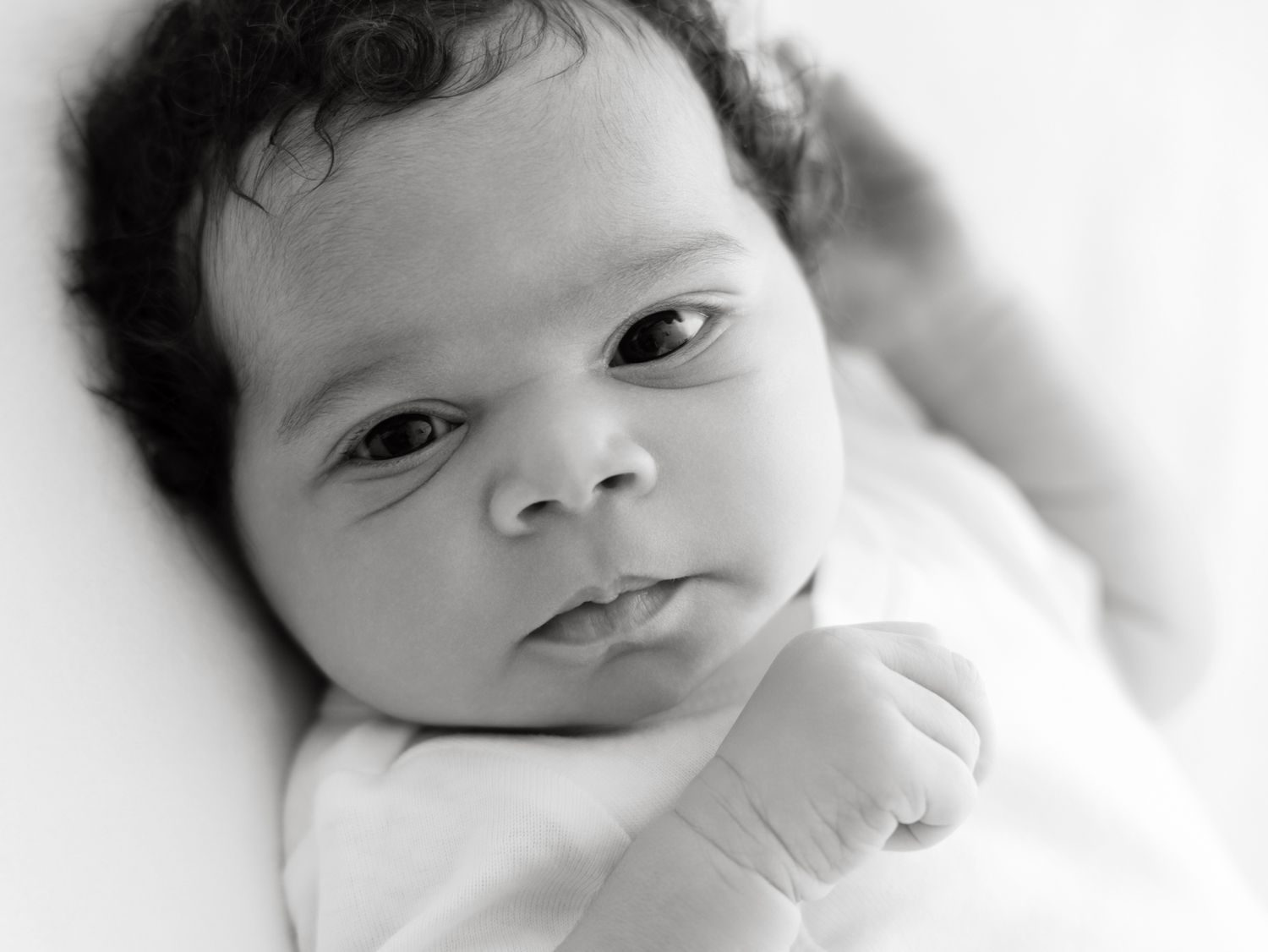Black and white portrait of a newborn baby with dark curly hair and alert eyes, lying on a white blanket.