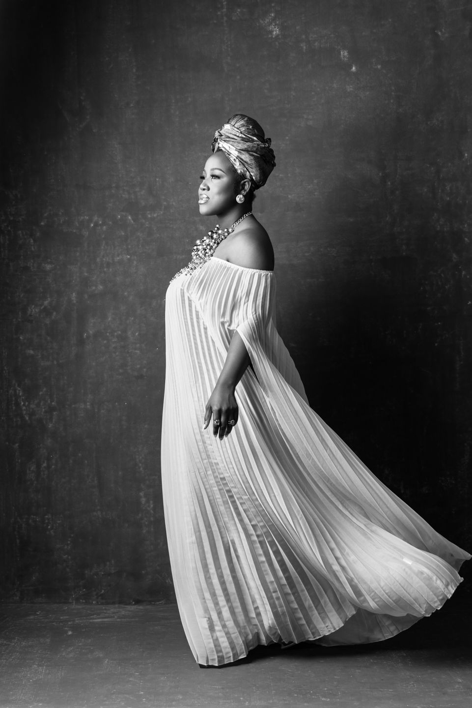 Black and white portrait of a woman in flowing white pleated gown and head wrap, posing gracefully in studio.