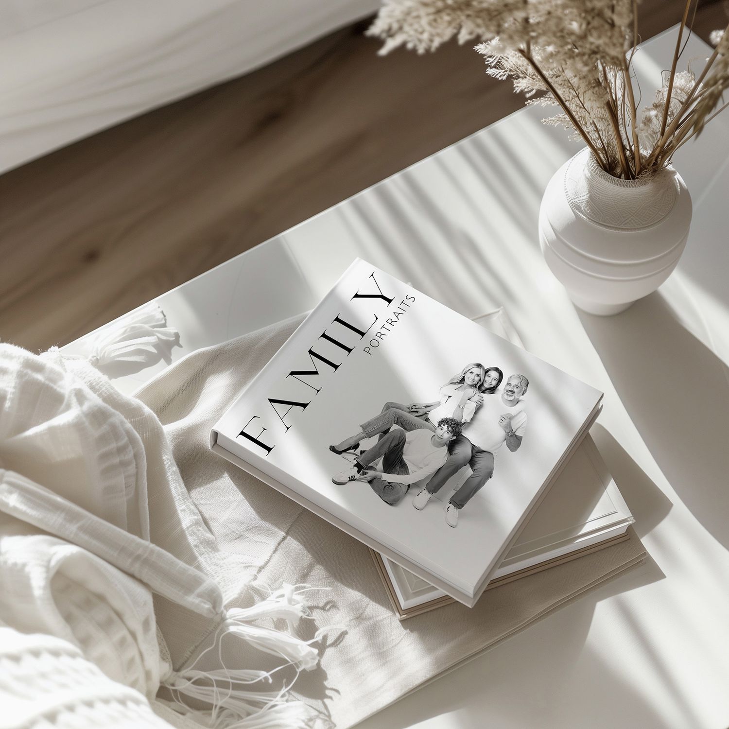 A white family photo book titled Family rests on a stack of books beside a white vase with dried flowers on a sunlit table.
