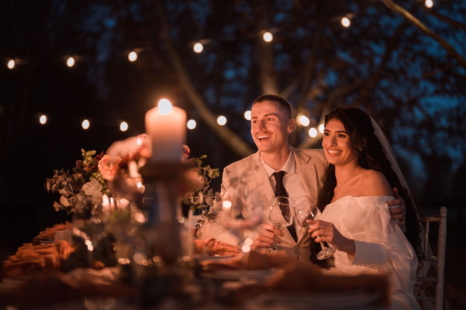 Romantic candlelit dinner scene with couple sharing laughter under twinkling outdoor lights.