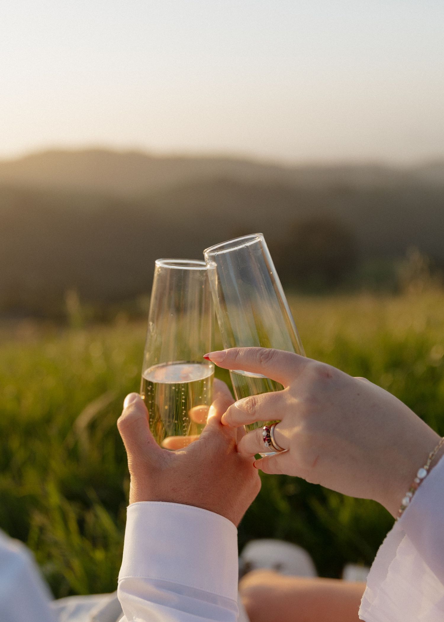 An engaged couple clink wine glasses together while enjoying a scenic sunset view over rolling hills at Prefumo Canyon in San Luis Obispo.