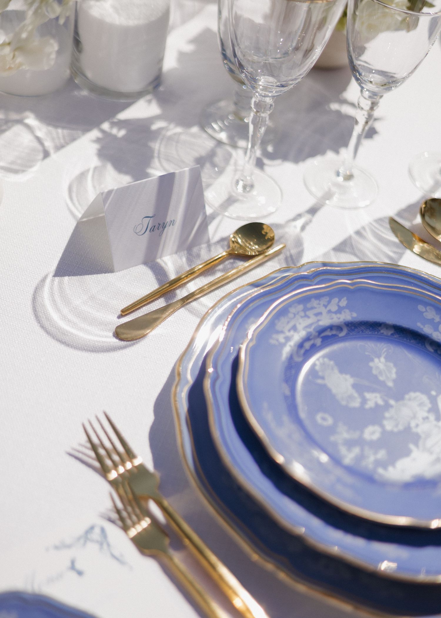 Blue and gold rimmed fine china place settings arranged on a white tablecloth for an elegant italian inspired wedding reception dinner.