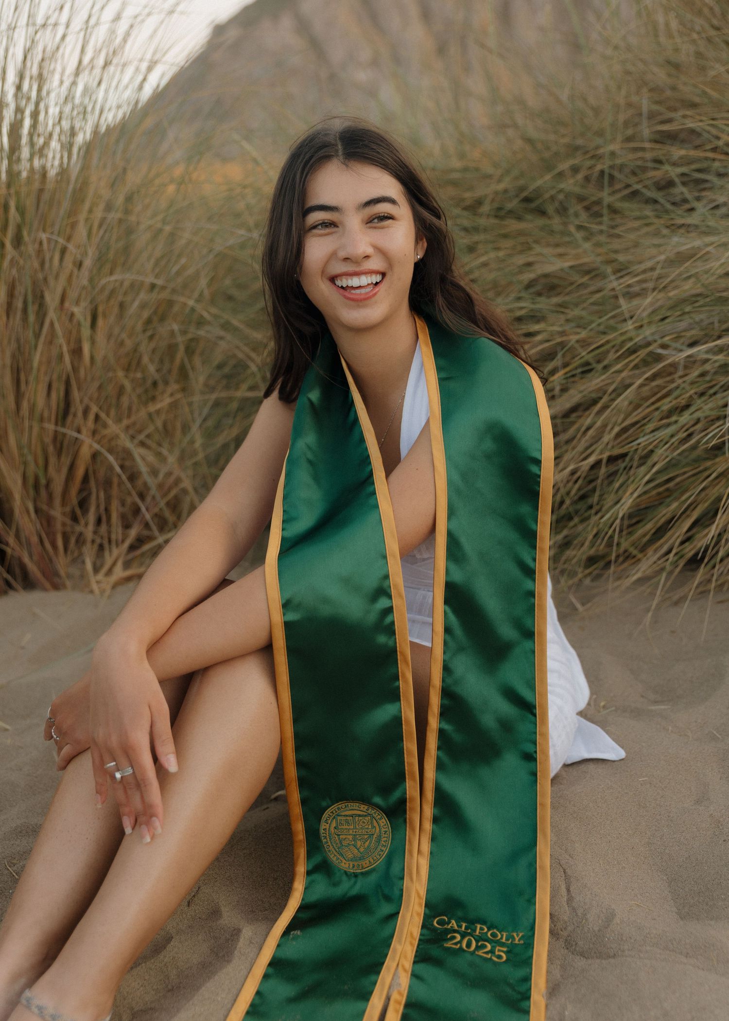 A Cal Poly San Luis Obispo graduate wearing her stole sits on sandy beach dunes during a golden hour photoshoot in Morro Bay, California.
