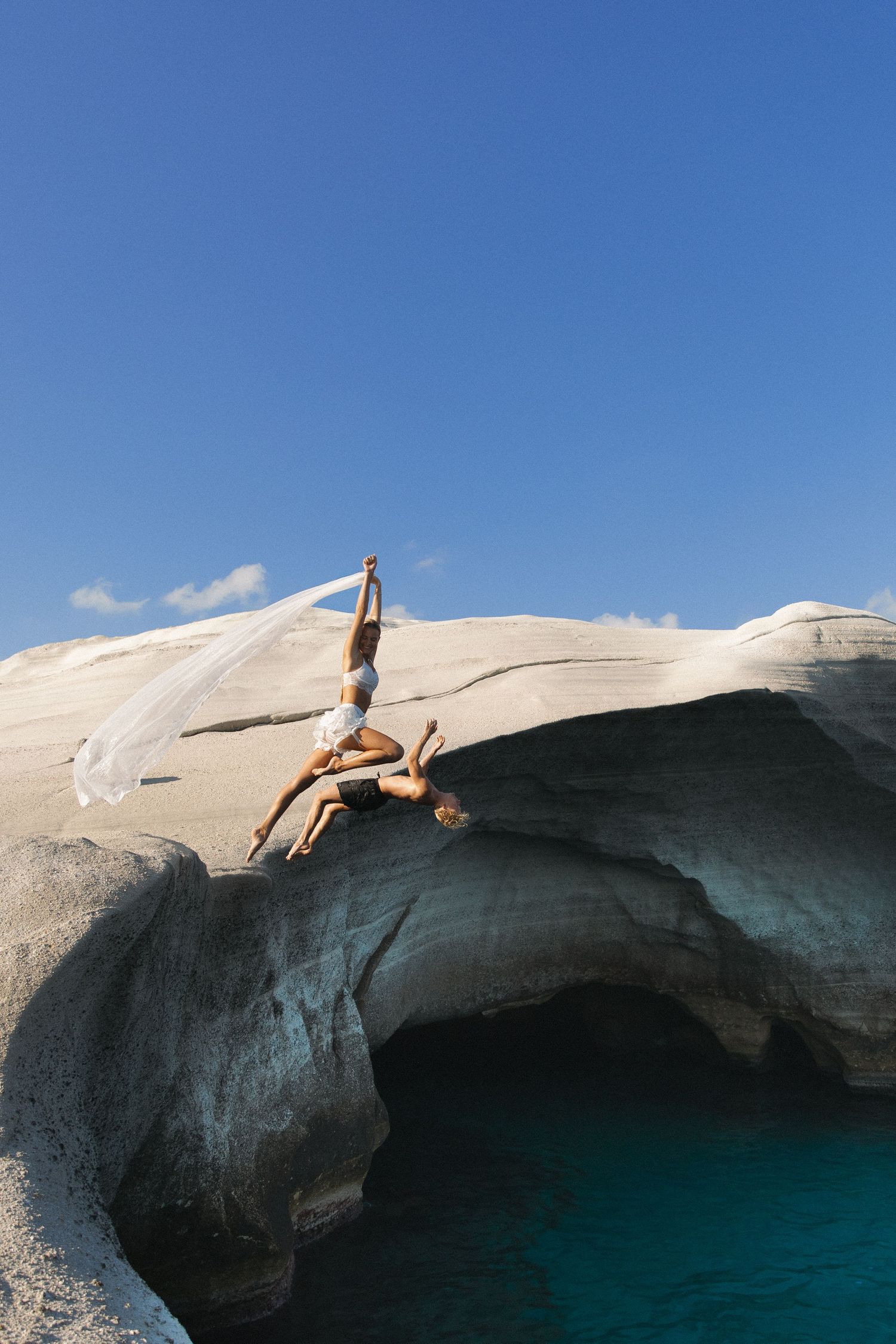 A newly married couple dives into crystal clear turquoise waters from white marble cliffs at Sarakiniko Beach in Milos, Greece.