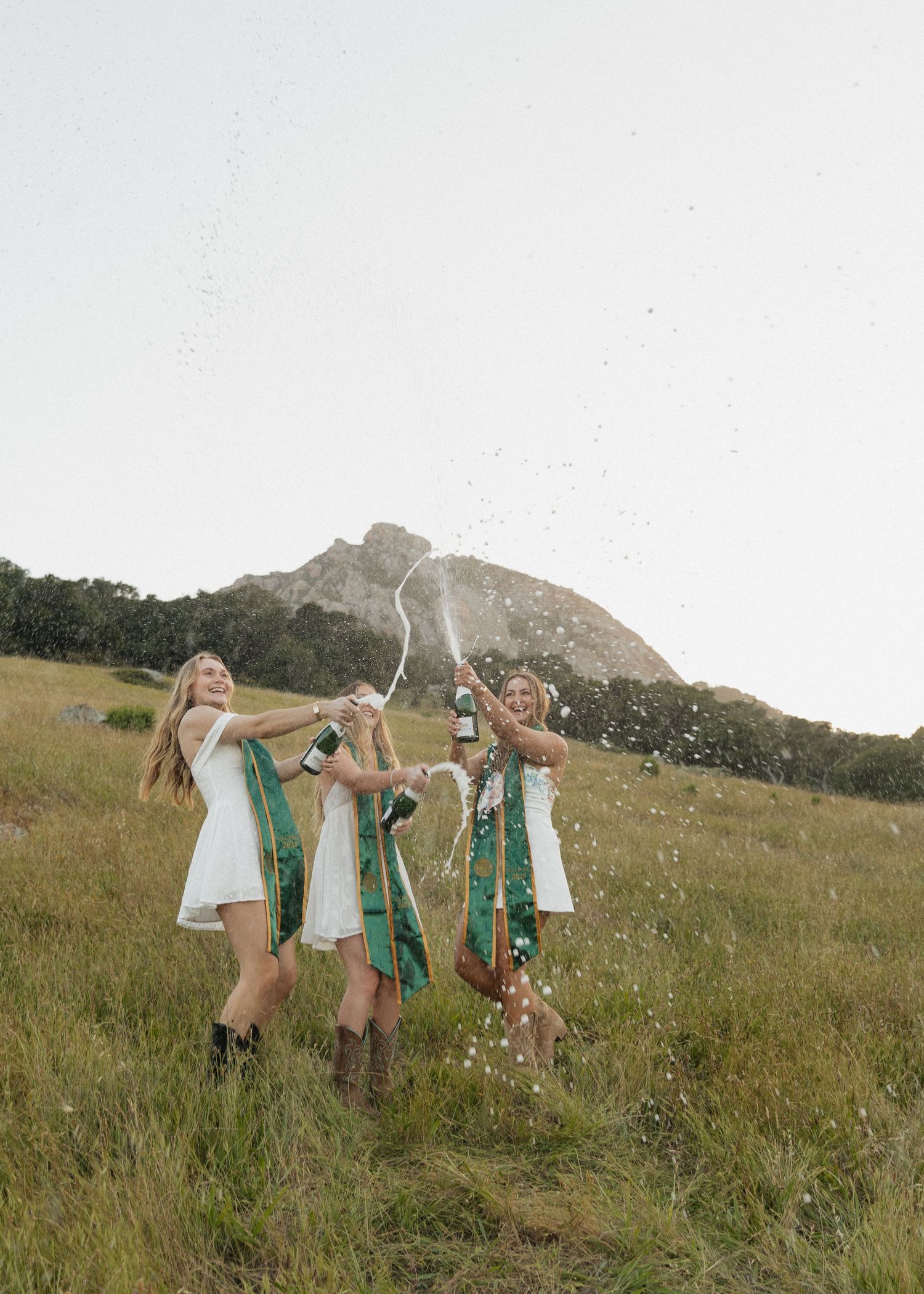 A group of three Cal Poly San Luis Obispo graduates celebrate joyfully by spraying champagne with Bishop's Peak in the background.