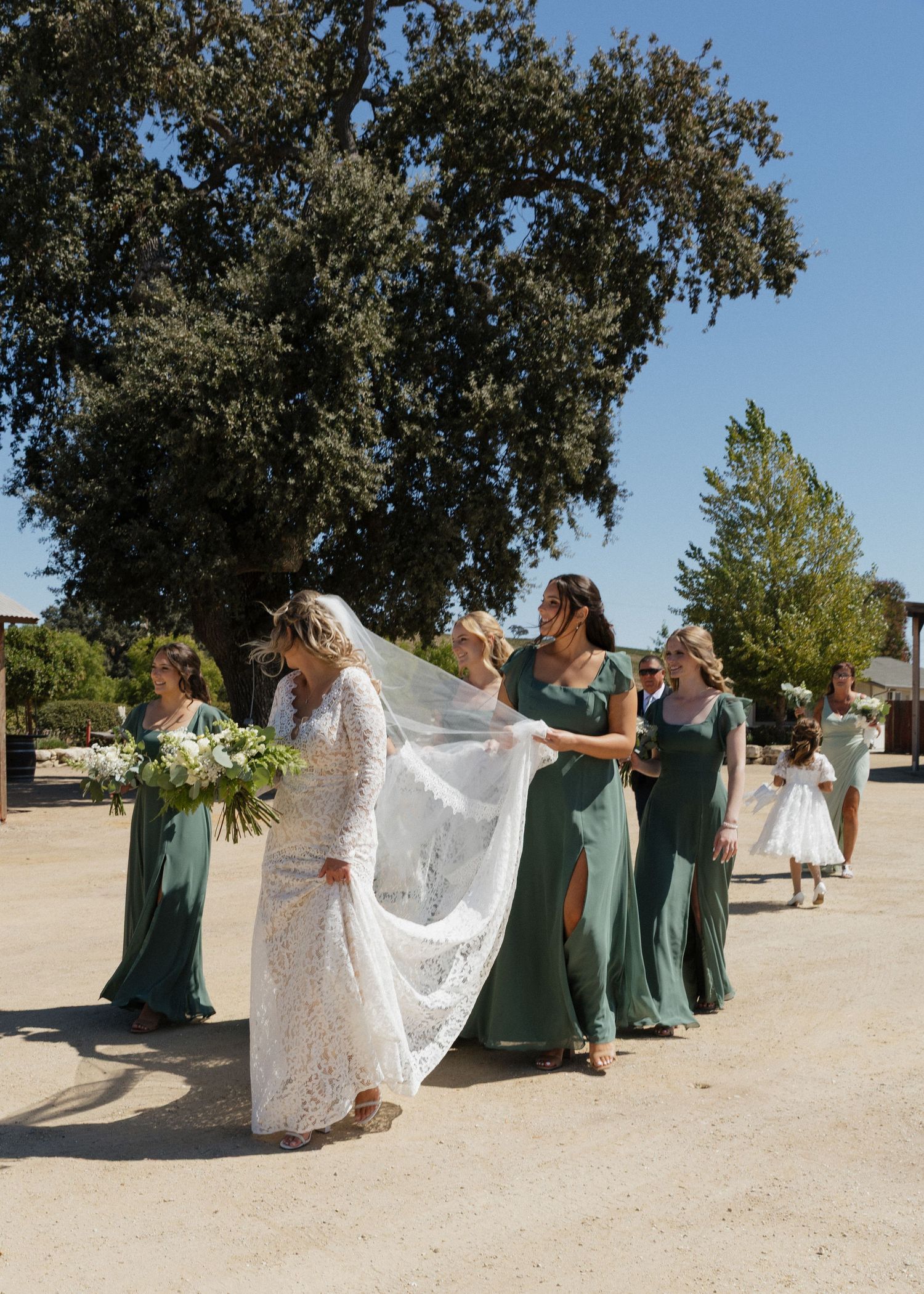 Bridesmaids in green dresses help hold up a bride's lace dress as they walk away from their venue in San Luis Obispo, California.