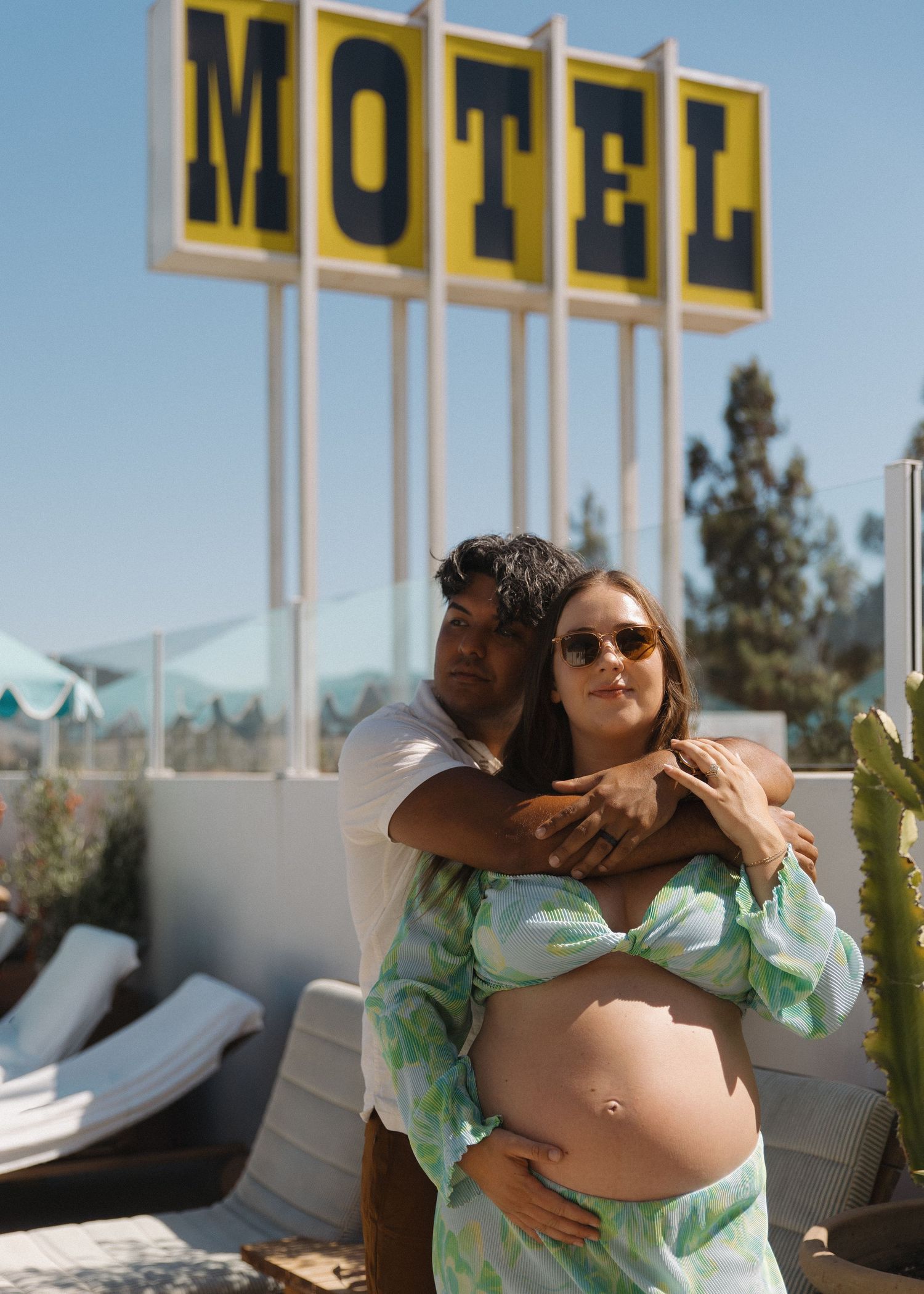 A pregnant couple hugging outside by the pool at the Skyview Motel in Los Alamos, California wearing 70s inspired attire.