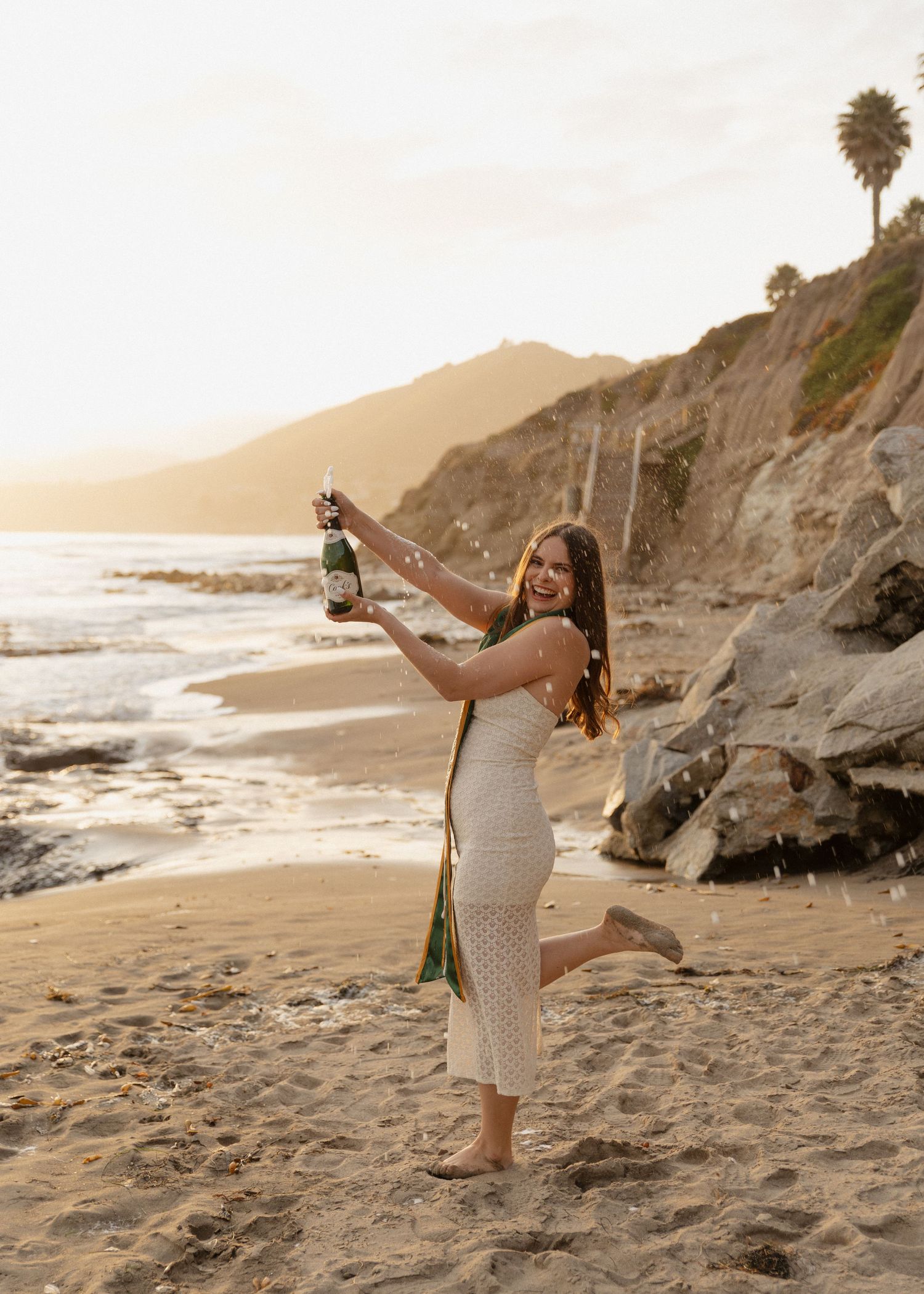 A Cal Poly San Luis Obispo graduate in a white dress pops champagne playfully on Shell Beach, California at sunset.