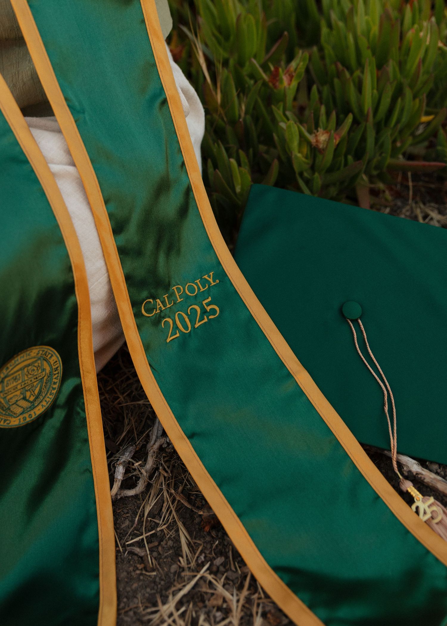 A green graduation gown with gold trim lies on grass, showing details of the academic regalia fabric and stitching.