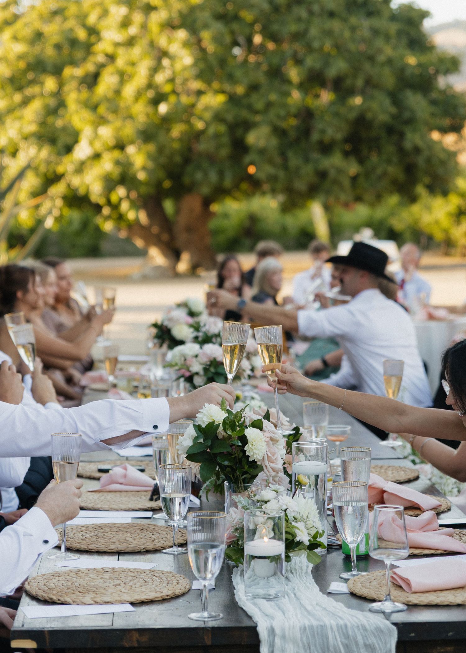 A wedding party cheers champagne glasses at an elegant outdoor reception dinner under golden evening light at Higuera Ranch in California.