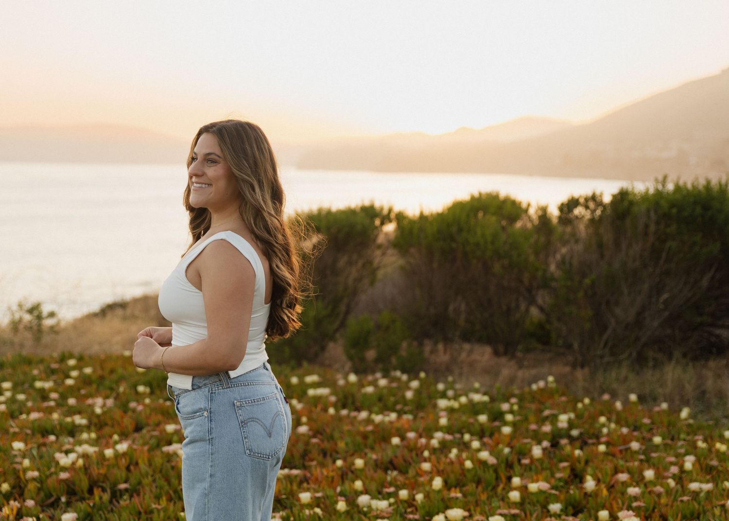 A Cal Poly SLO graduate stands in a wildflower meadow in Shell Beach, California at golden hour with mountains in the background.