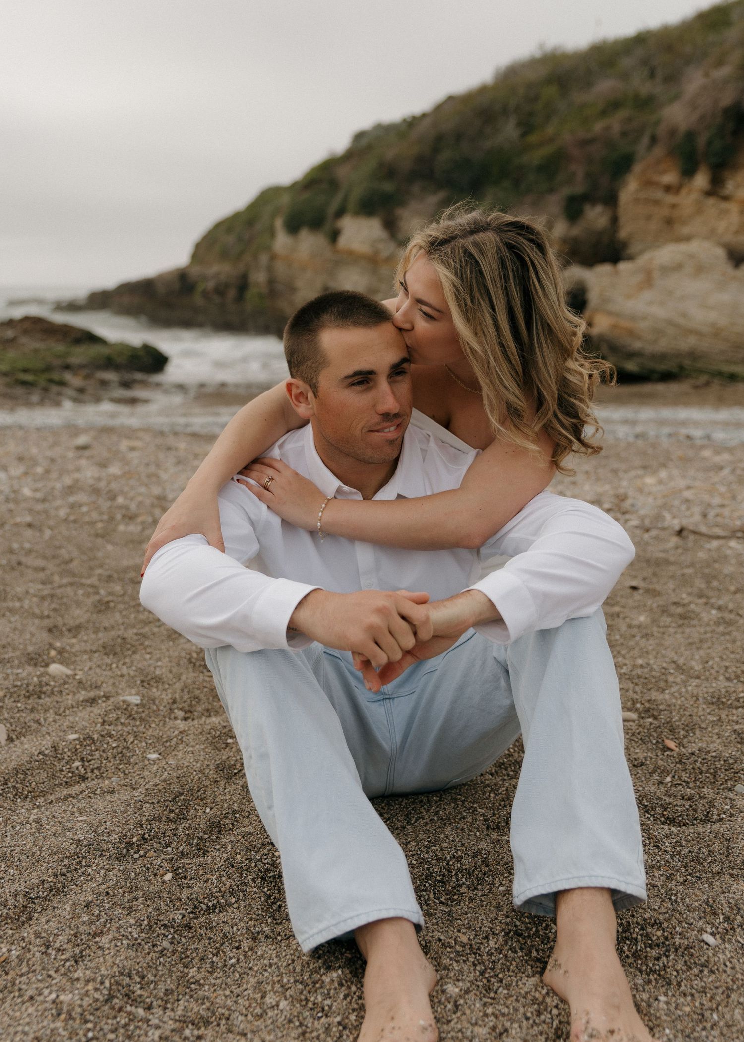 A couple in white clothing sits intimately together on a rocky beach at Montana De Oro Beach in Los Osos, California.