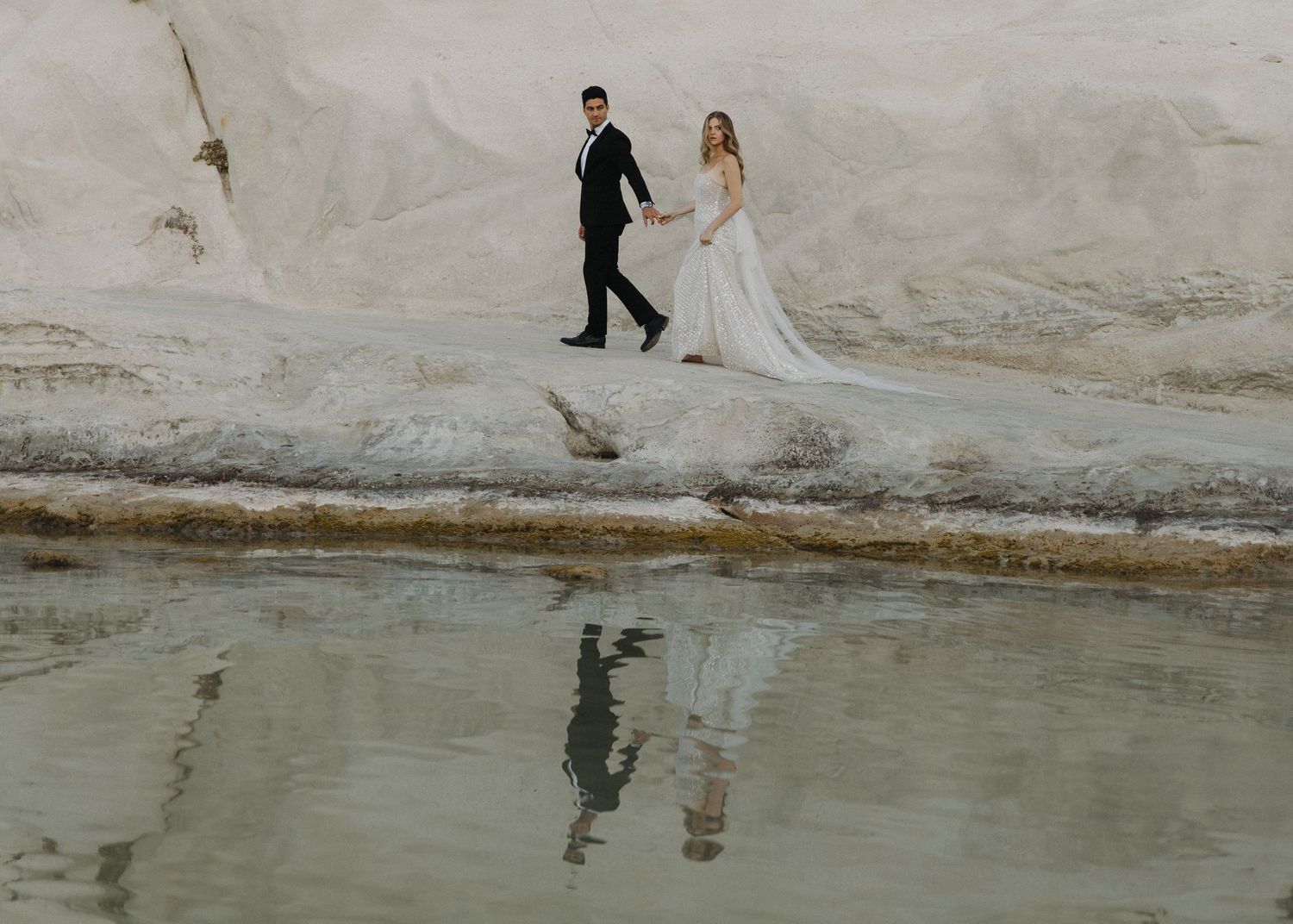 A couple in wedding attire walks along a rocky beach shoreline in Milos, Greece, creating romantic silhouettes against the water.
