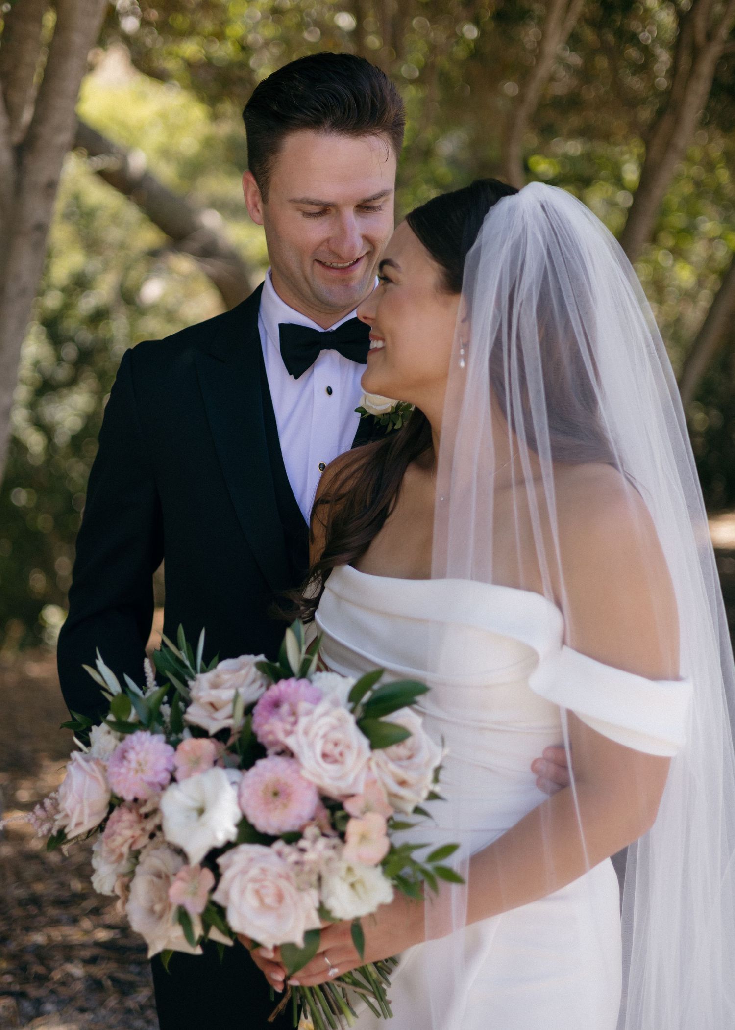A romantic bride and groom embraces in the olive grove at Higuera Ranch in San Luis Obispo, California holding a soft pink floral bouquet.