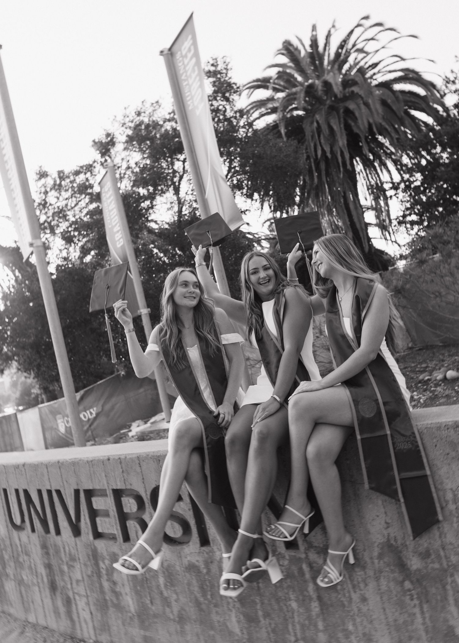 A group of Cal Poly San Luis Obispo graduates sitting on the campus entrance sign holding up their caps.