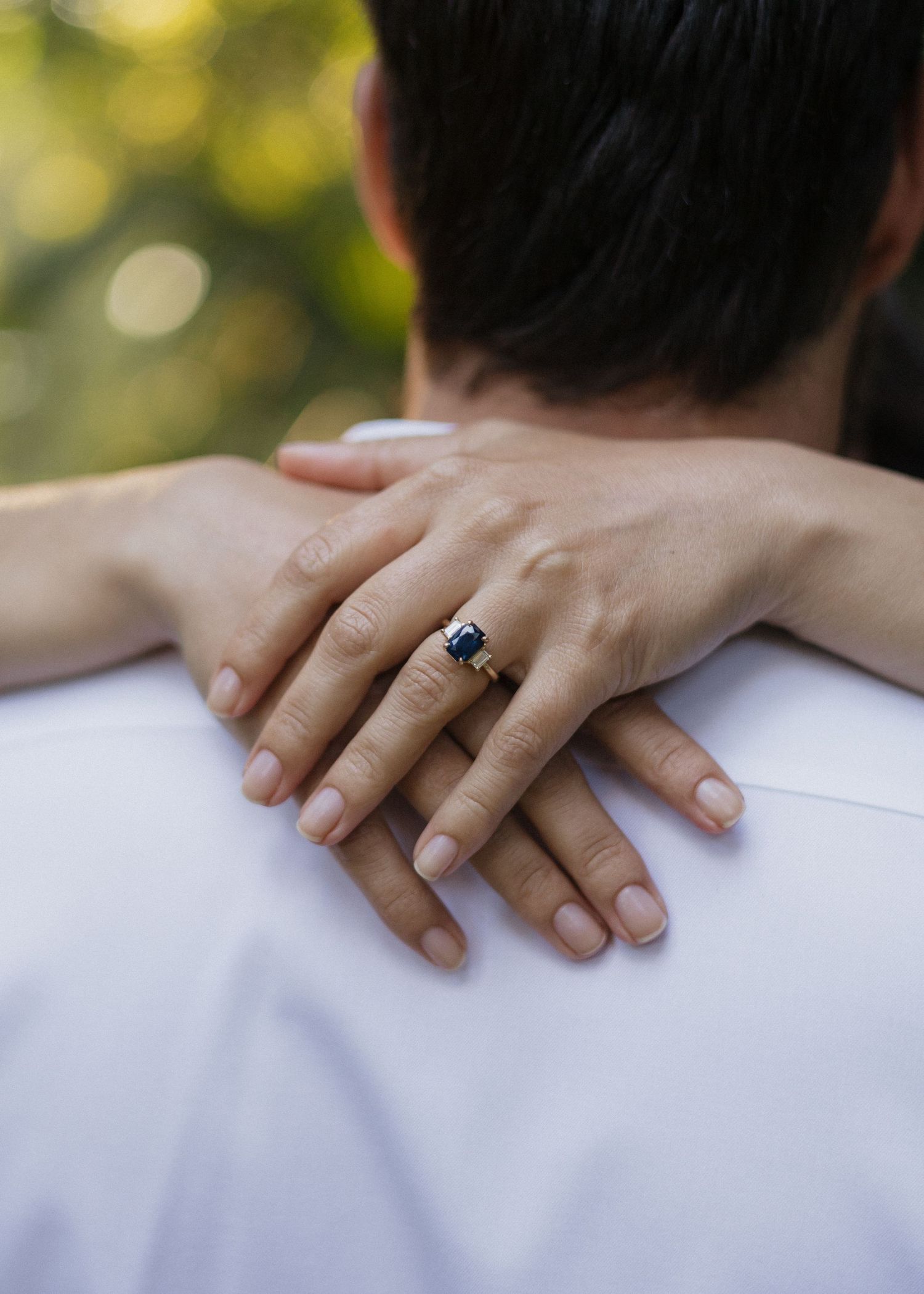 Close-up of hands wearing an engagement ring with a blue gemstone resting on fiances shoulders in natural light.