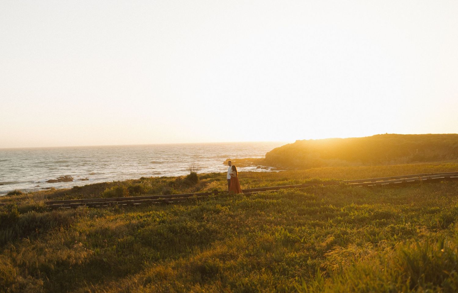 A couple walking at Moonstone Beach boardwalk in Cambria, California with scenic sunset over the Pacific Ocean.