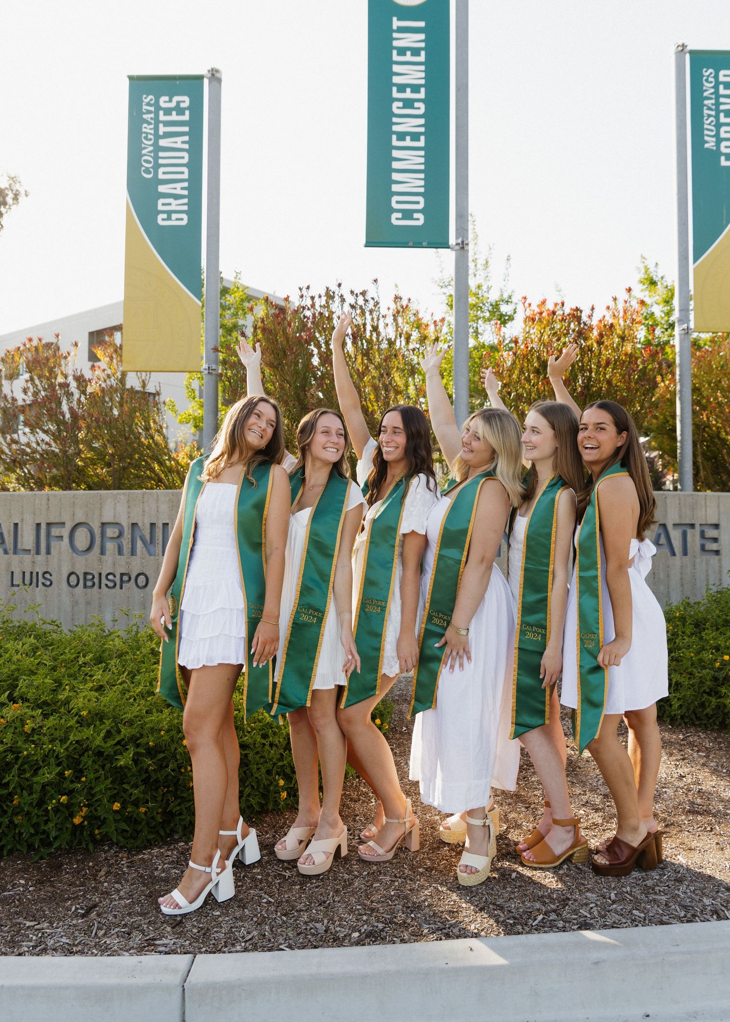 A group of Cal Poly San Luis Obispo graduates standing, laughing at each other in front of the campus sign at sunset.