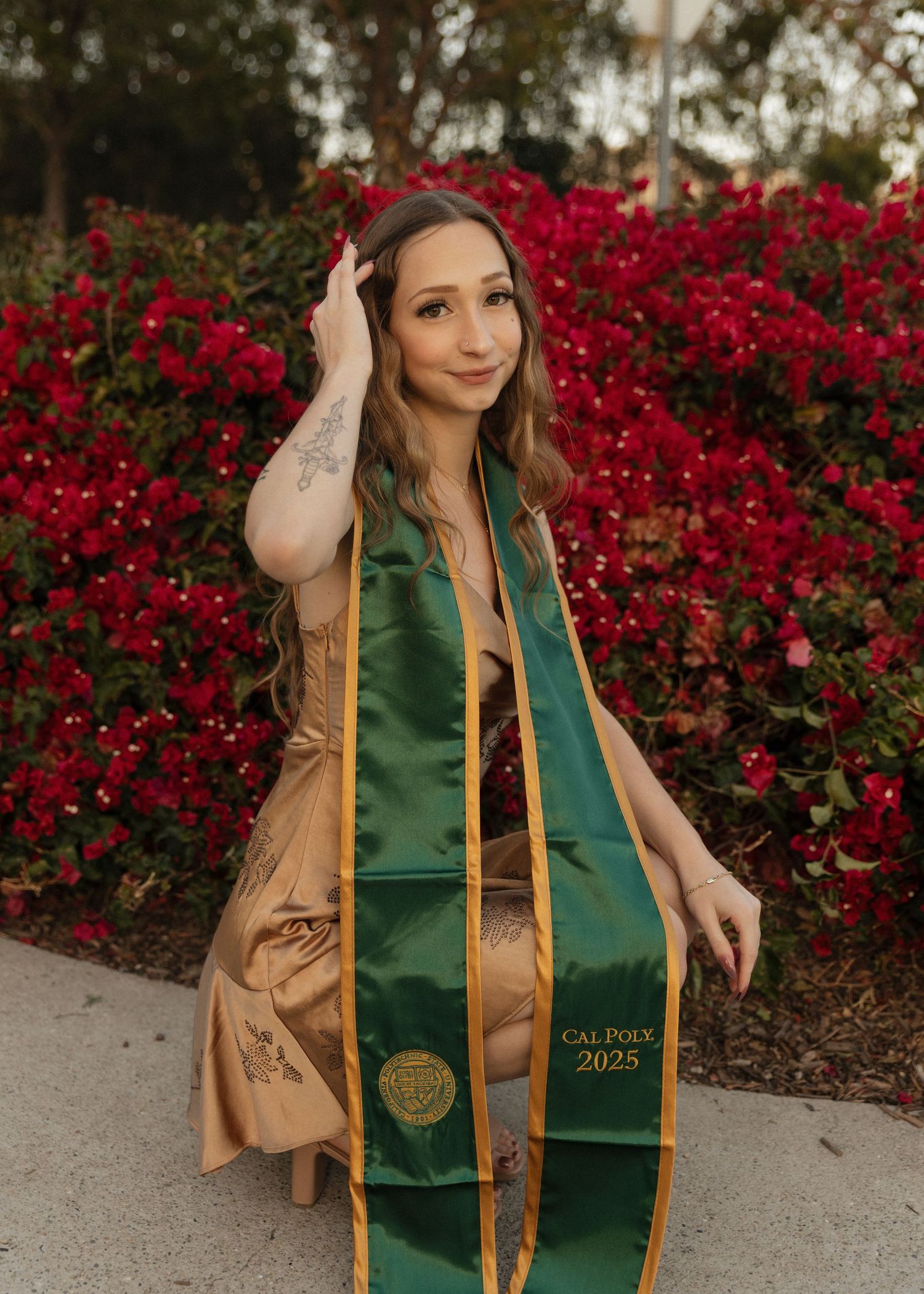 A Cal Poly San Luis Obispo graduate wearing her stole poses in front of vibrant red bougainvillea flowers during sunset.