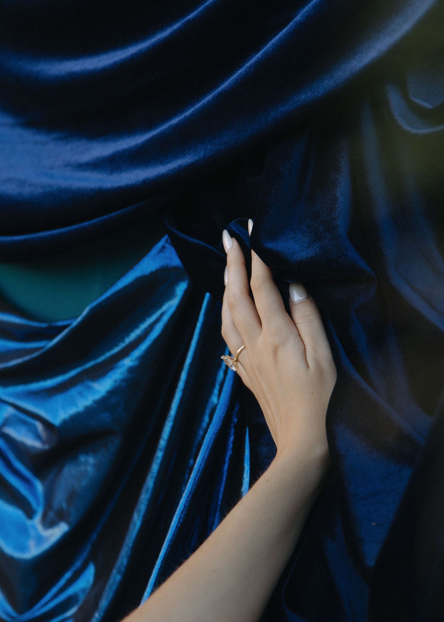 A hand wearing a gold engagement ring gently caresses a silky dark blue fabric creating elegant folds and shadows.