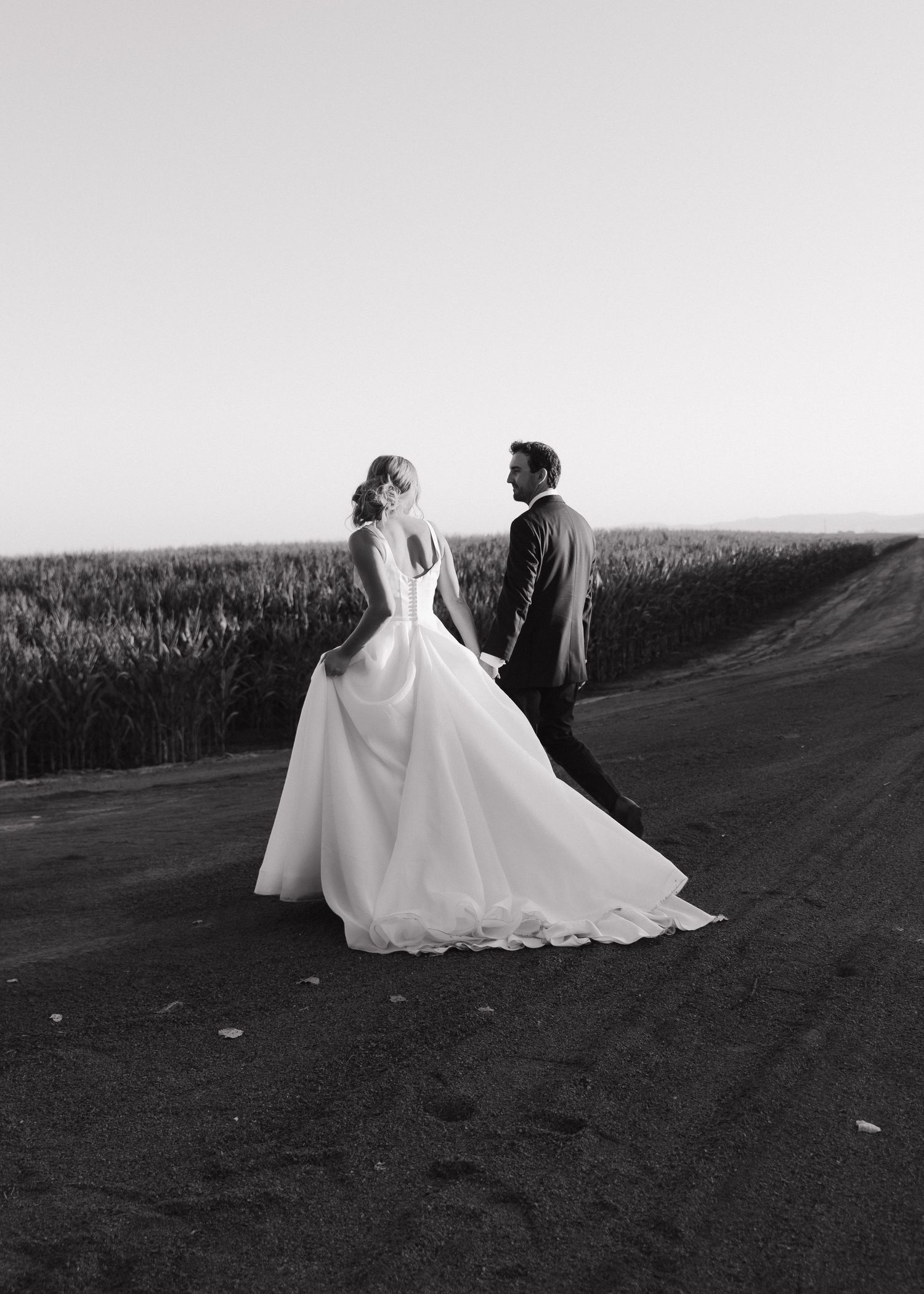 A black and white wedding photo showing a bride and groom walking on a dirt path next to a grassy field at sunset.