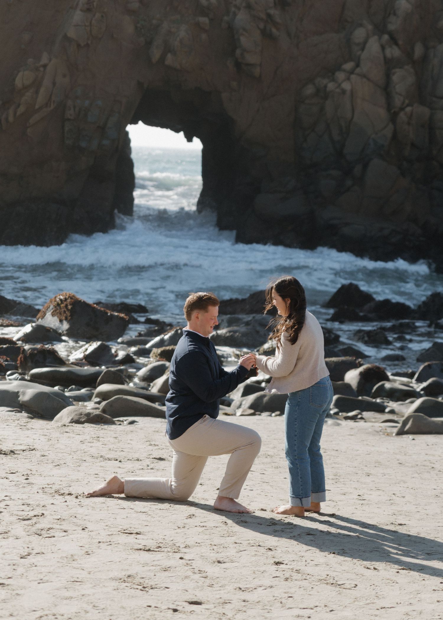 A man down on one knee proposing to a woman on a rocky beach with dramatic cliffs and ocean waves in the background in Big Sur, California.