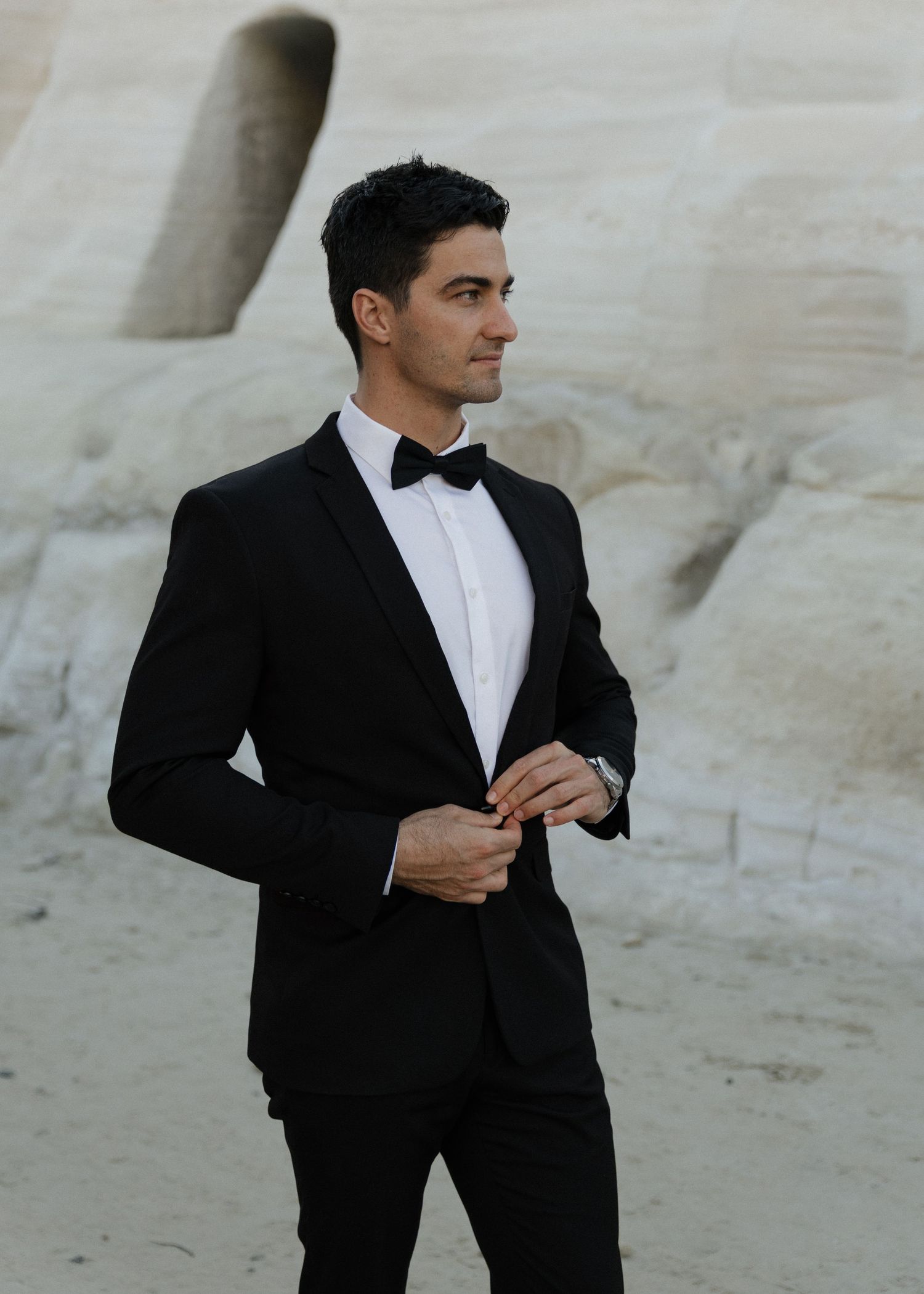 A groom in a classic black tuxedo and bowtie poses against Sarakiniko Beaches white stone backdrop in Milos, Greece.