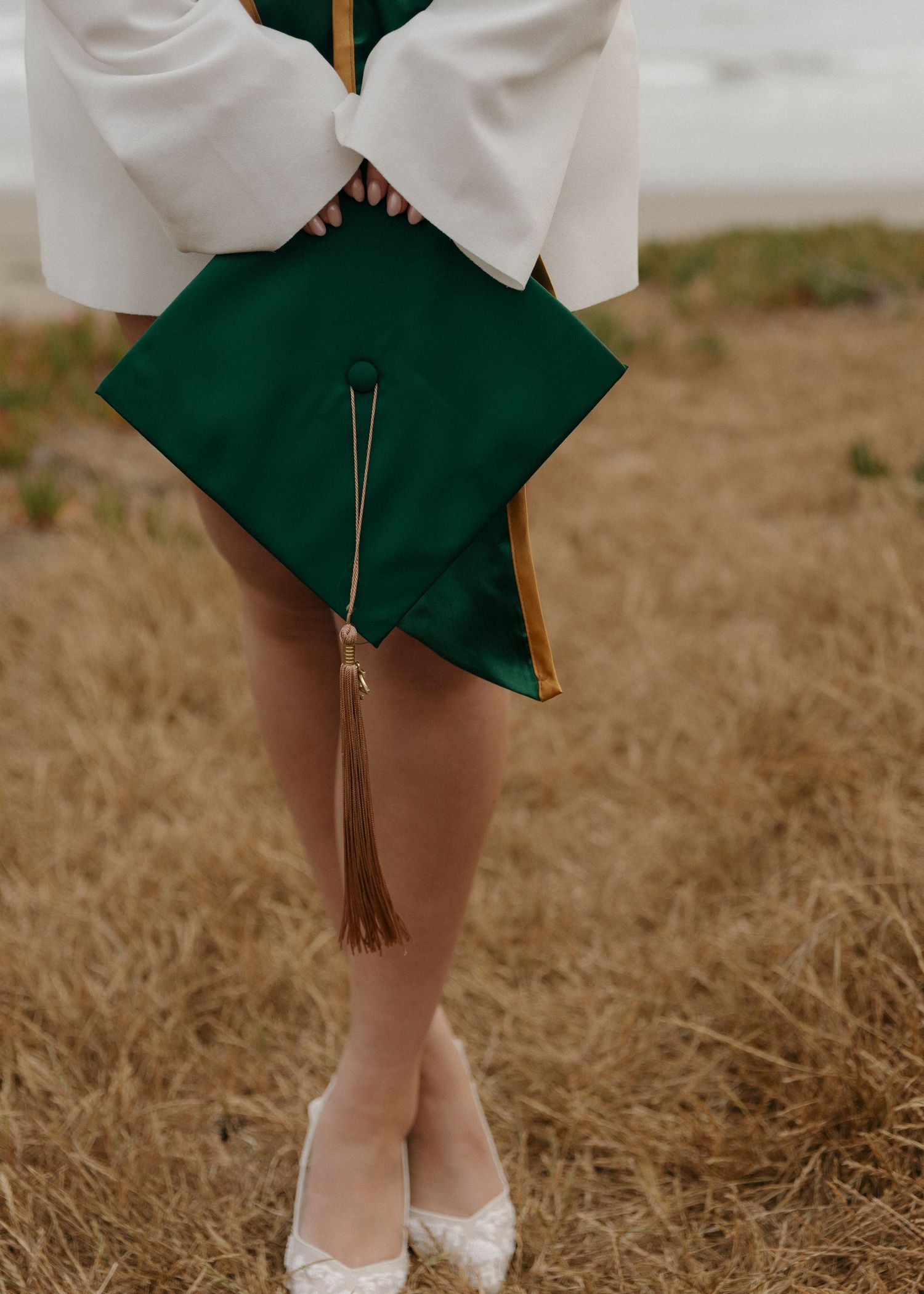 A detail photo of a Cal Poly San Luis Obispo graduate in white tiffany heels holds green cap cap by her legs.