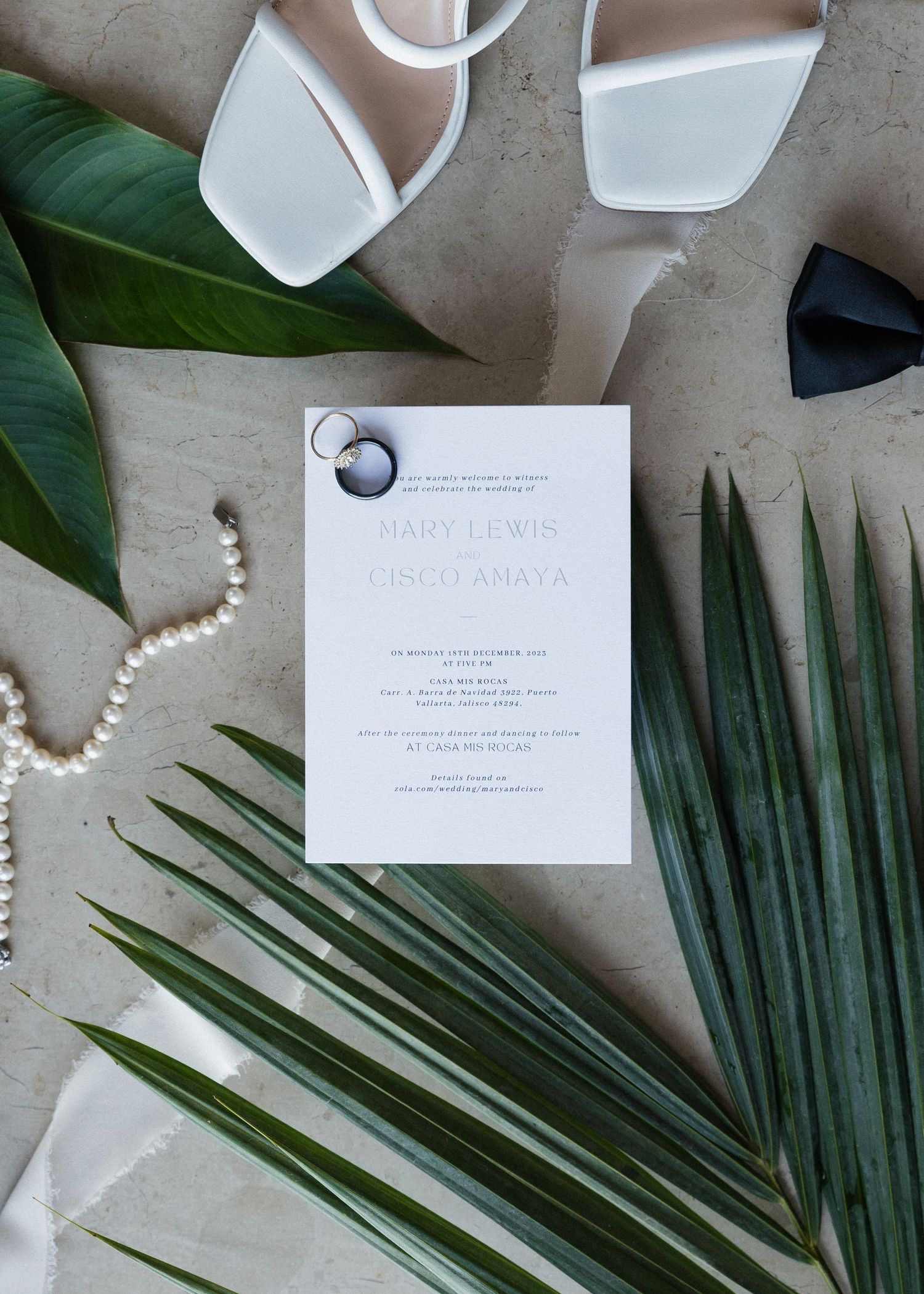 Elegant wedding invitation flatlay with palm leaves, pearls, and white stationery on a neutral textured background.