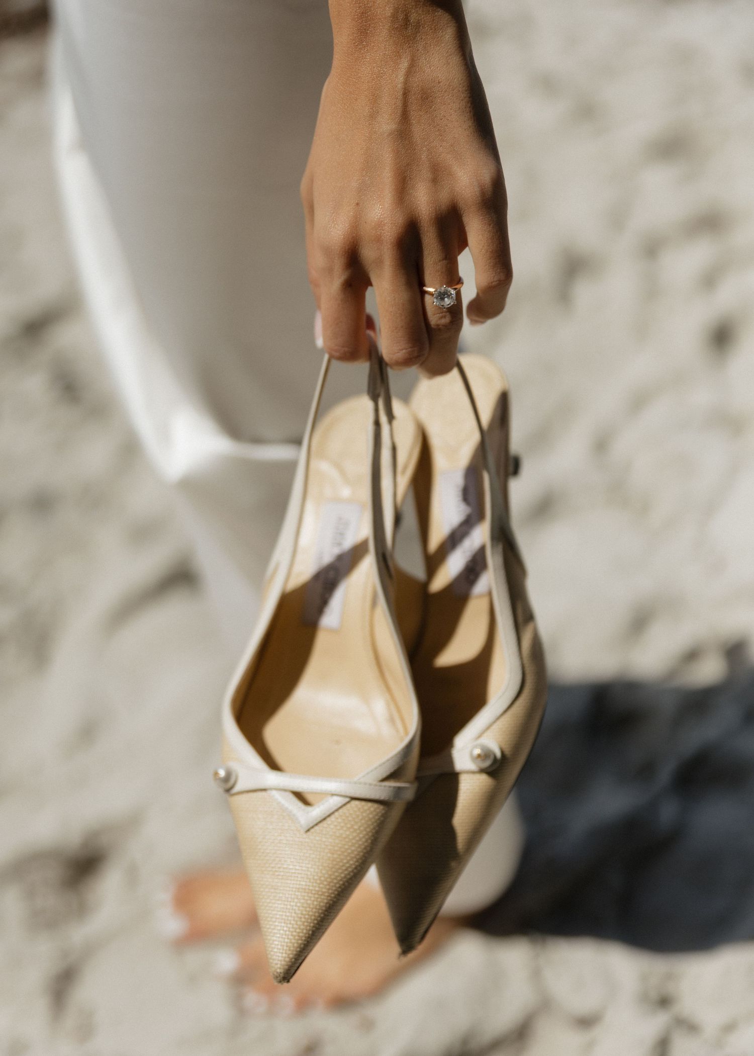 A brides hand holds a pair of elegant beige kitten heel slingback shoes against a sandy beach background.