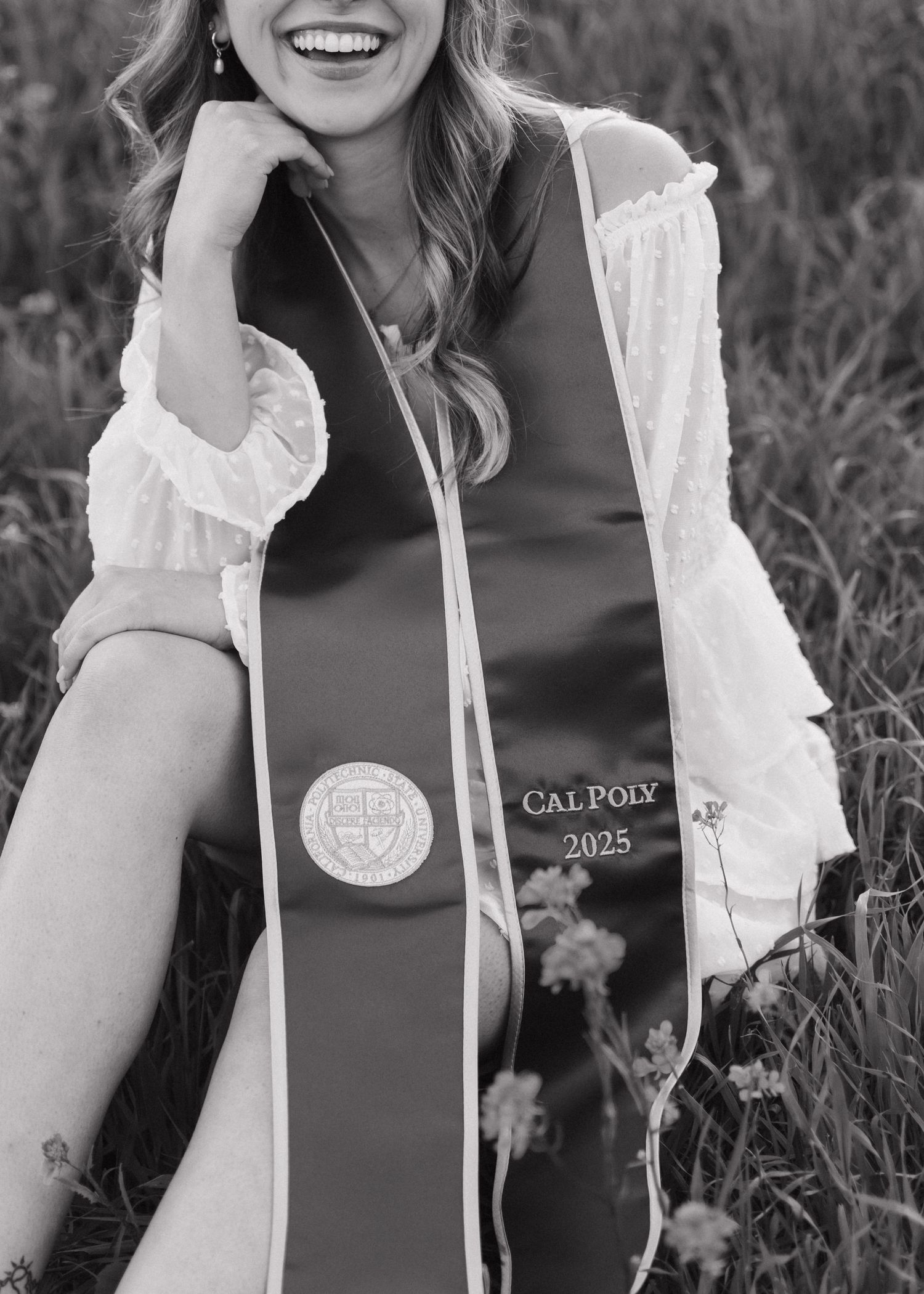A black and white photo of a graduate wearing a Cal Poly stole smiles during commencement celebrations at Bishops Peak in San Luis Obispo.