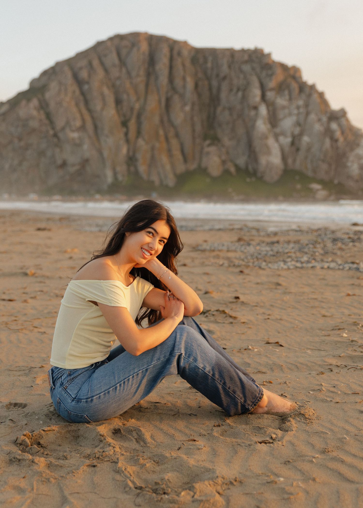 A graduate sits on a sandy beach in California at sunset with dramatic Morro Bay Rock in the background.