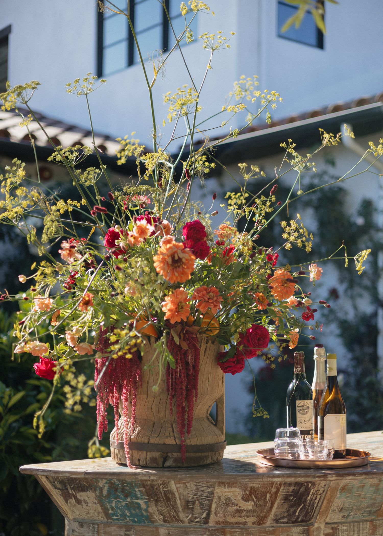 Rustic floral arrangement with red and orange blooms in vintage container on weathered table with wine bottles for a wedding bar setup.