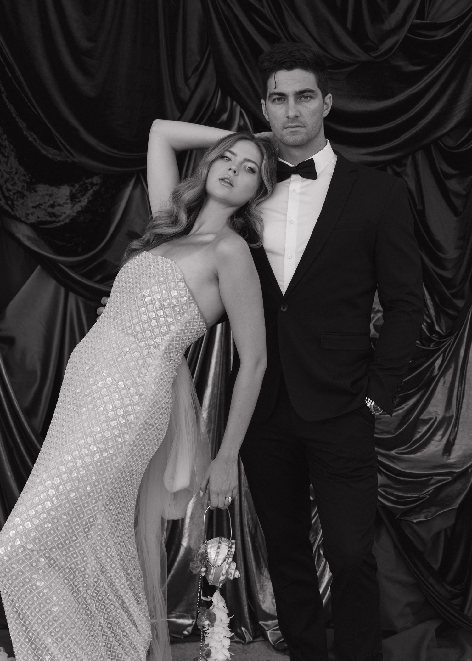 Glamorous bride and groom pose against dark blues draped backdrop in black and white portrait in Milos, Greece.