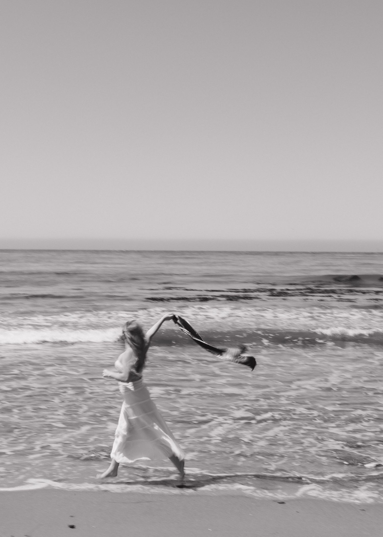 Black and white photo of a Cal Poly San Luis Obispo graduate in white dress holding her stole along a beach shoreline in Shell Beach.