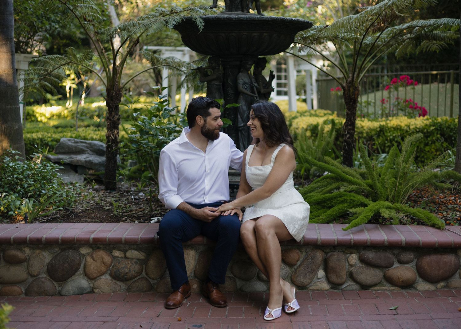 A couple sits together on a stone wall in a lush garden setting at Hartley Botanica in Camarillo, California for a romantic moment.
