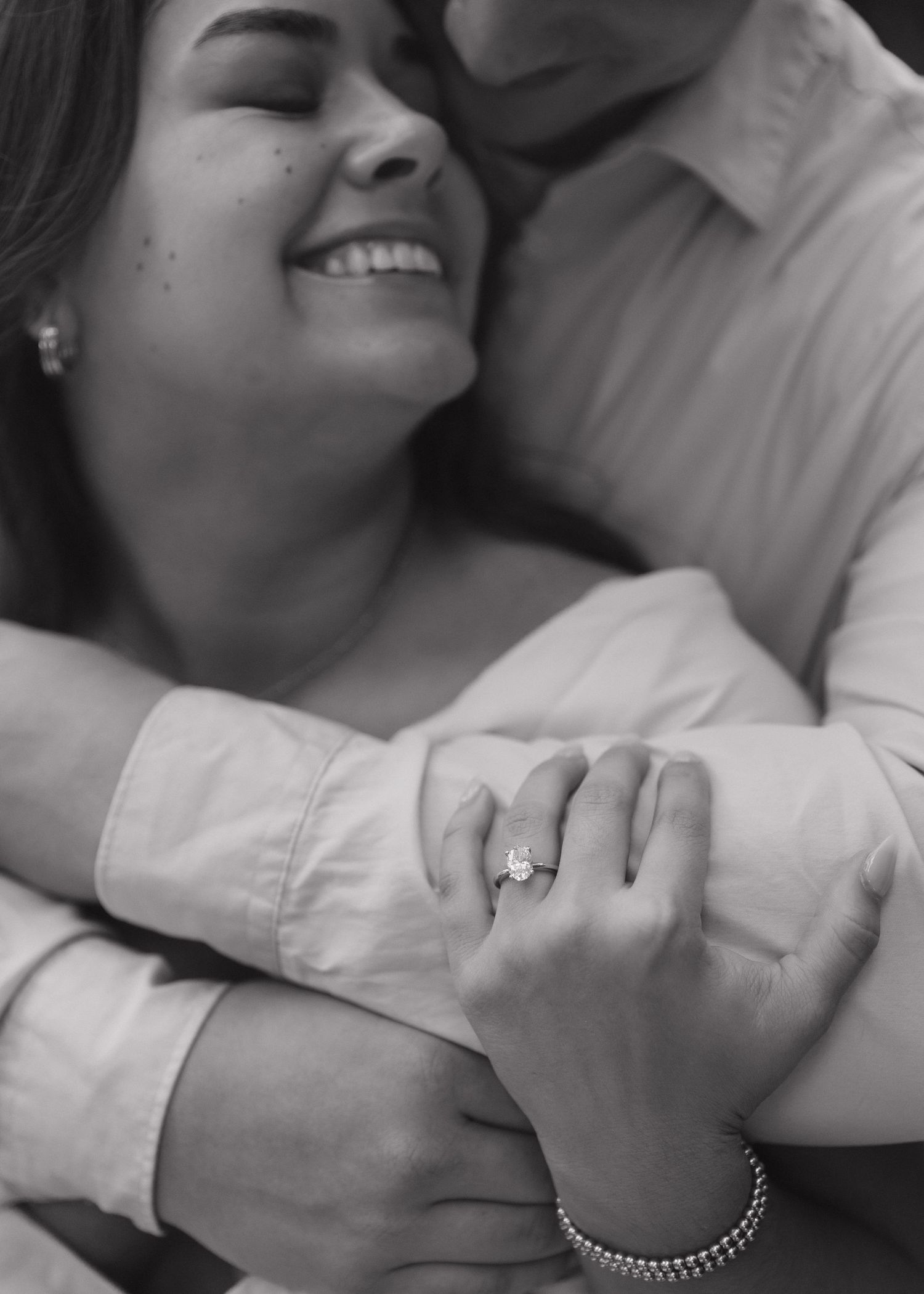 A black and white photo of a newly engaged couple sharing an intimate hug with hands clasped and showcasing the engagement ring.
