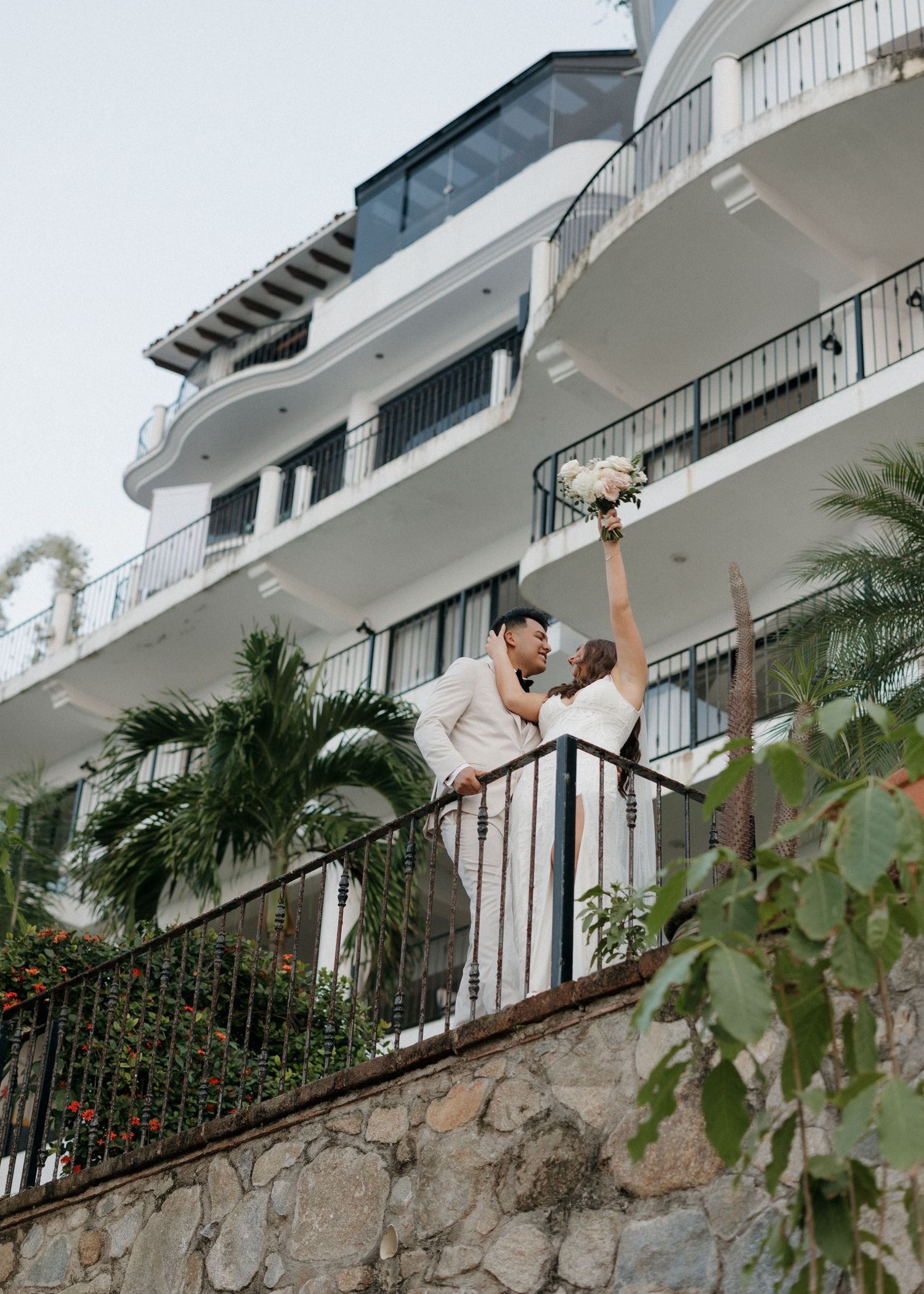 Bride and groom stand on a balcony raising her bouquet after their intimate wedding ceremony of the coast of Puerto Vallarta, Mexico.