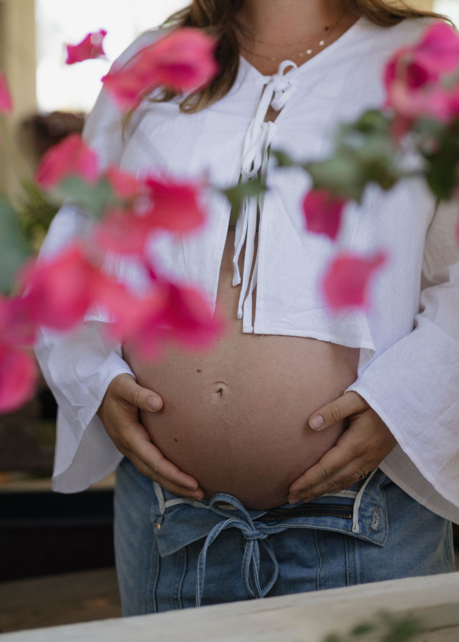 Close-up view of a pregnant belly wearing a white top and blue jeans with pink flowers visible in the background.