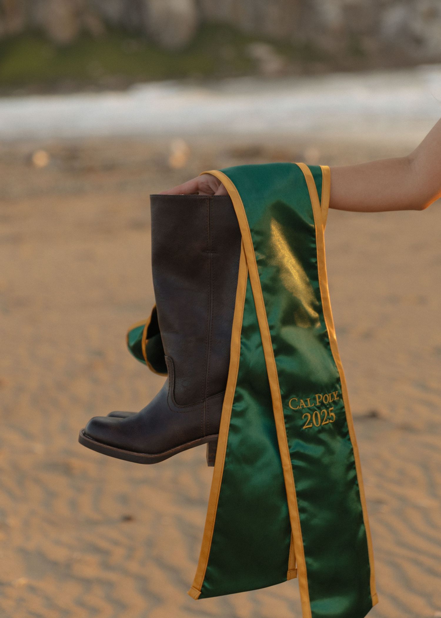 A detail photo of a Cal Poly San Luis Obispo graduate holding her boots and stole with Morro Bay beach in the background.
