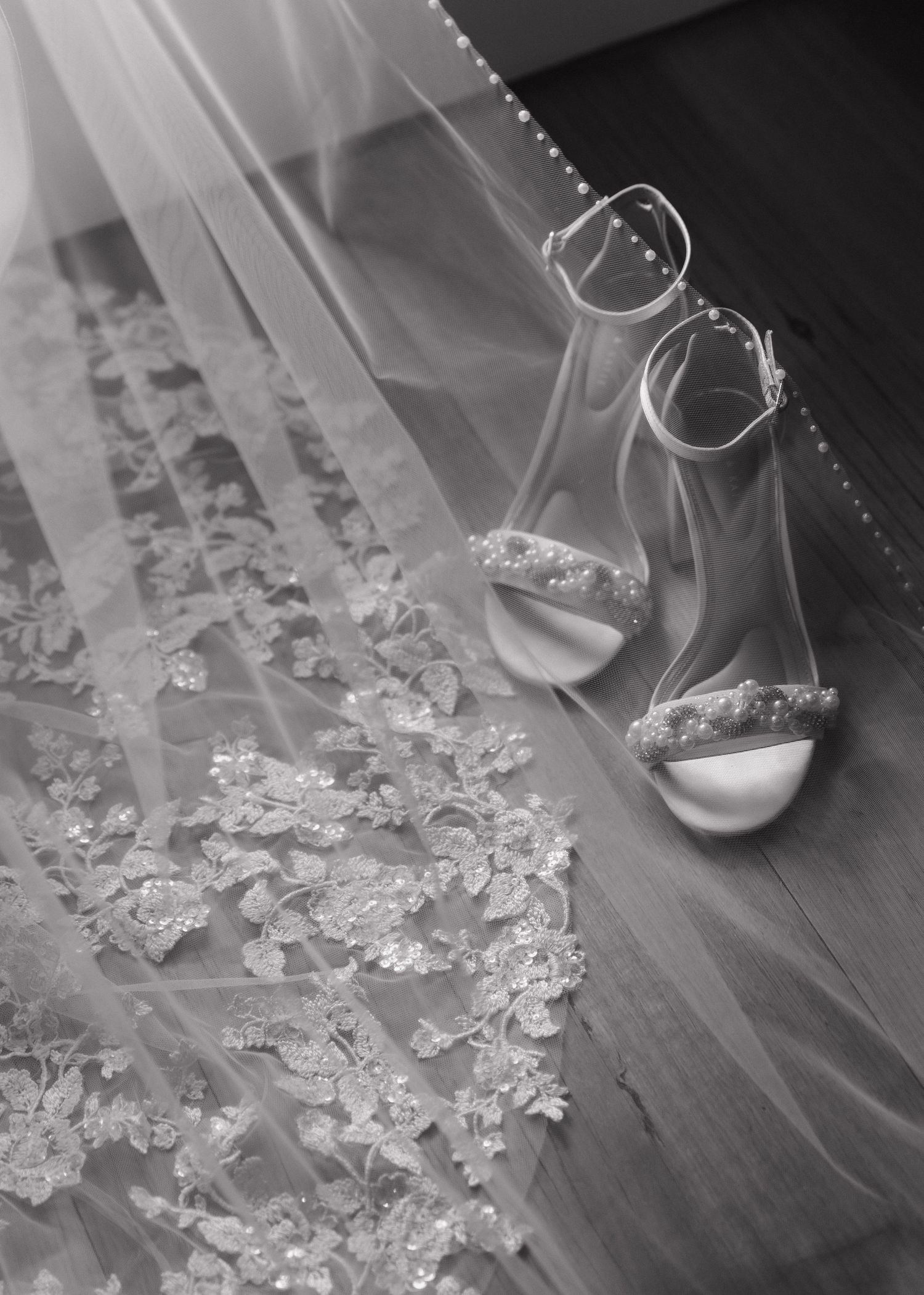 A white high-heeled bridal shoe stands elegantly on a wooden floor surrounded by scattered lace and fabric petals.