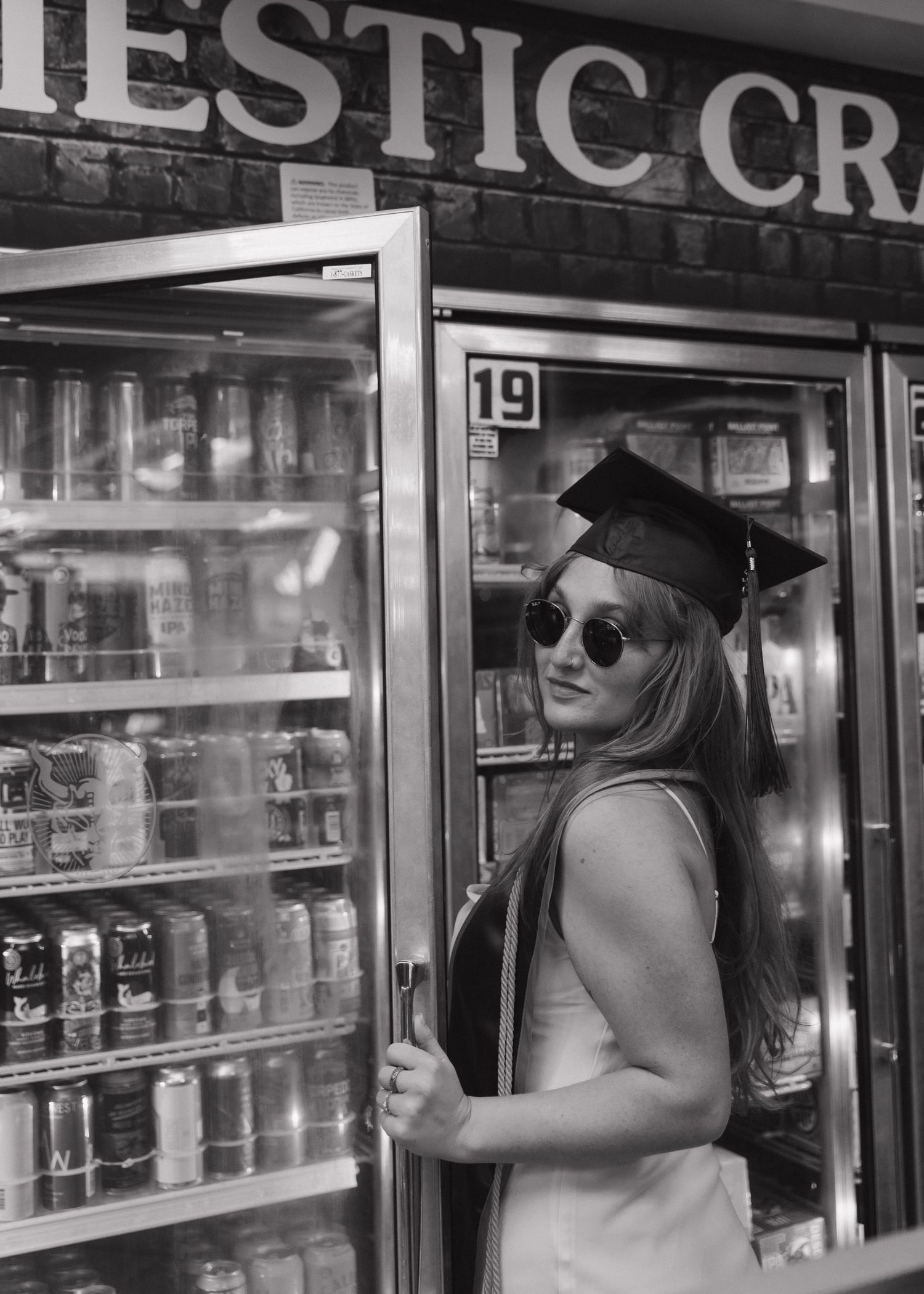 A Cal Poly San Luis Obispo graduate in a cap and sunglasses poses playfully in front of Sandy's Liquor Store refrigerator doors.