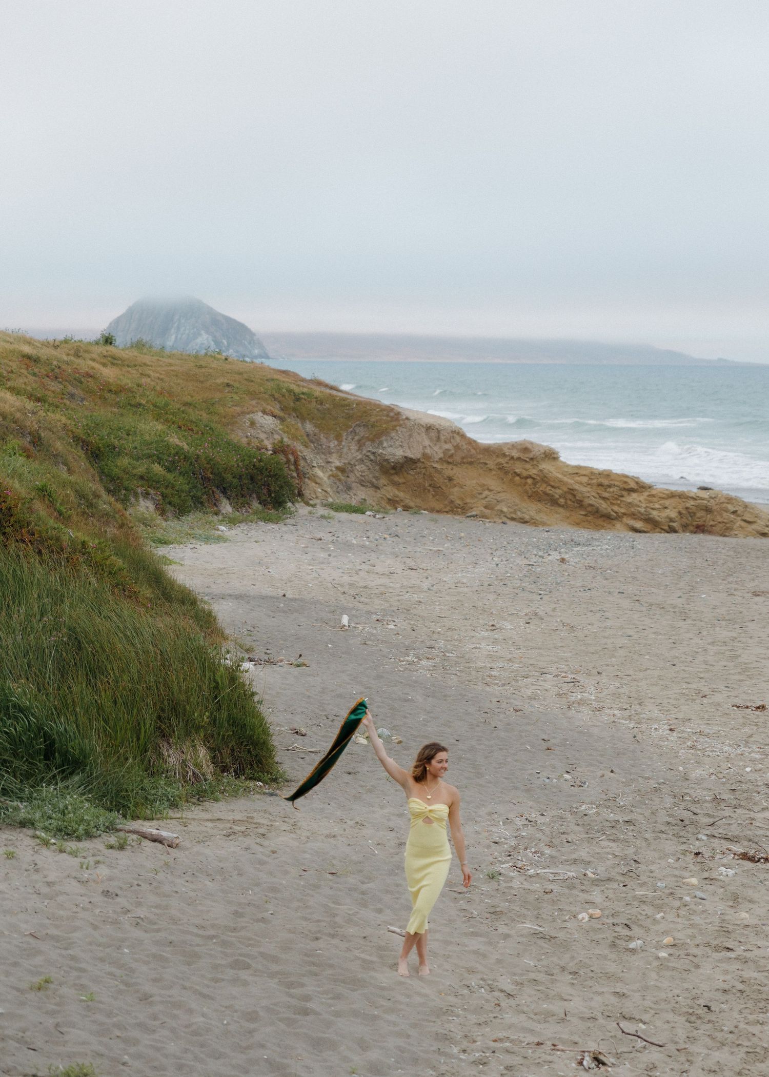 A photo showing a Cal Poly San Luis Obispo graduate in a yellow dress walking down a misty beach with Morro Bay rock in the background.