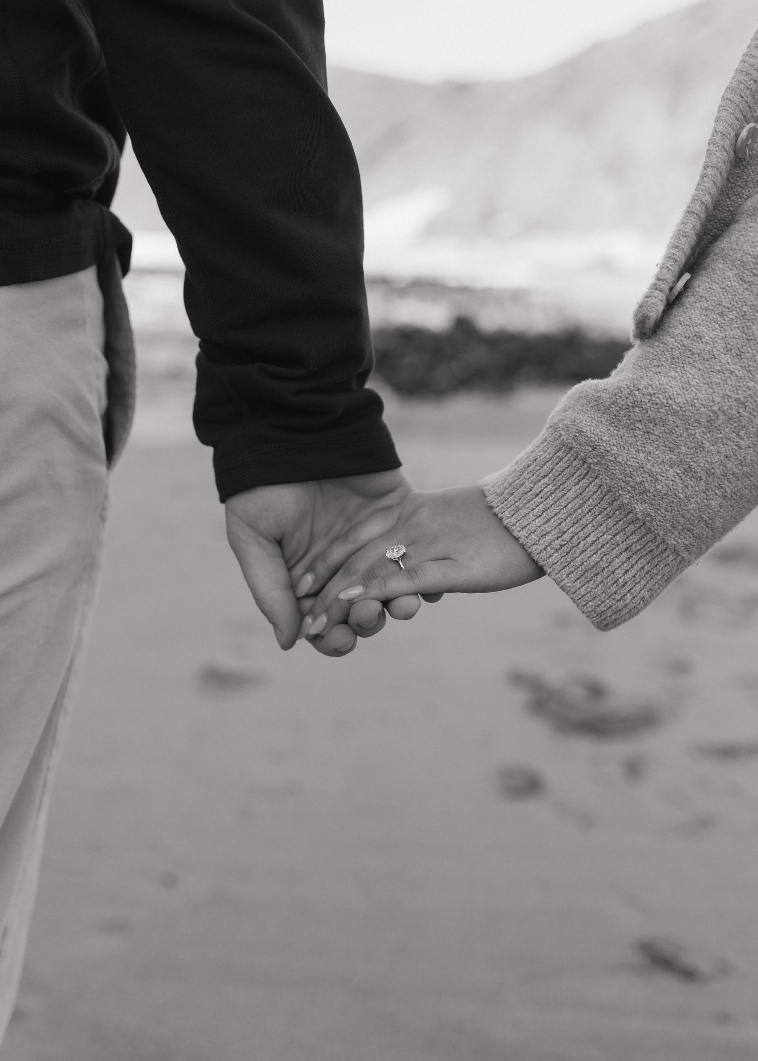 A black and white close-up of a newly engaged couple holding hands with a blurred Big Sur, California in background.