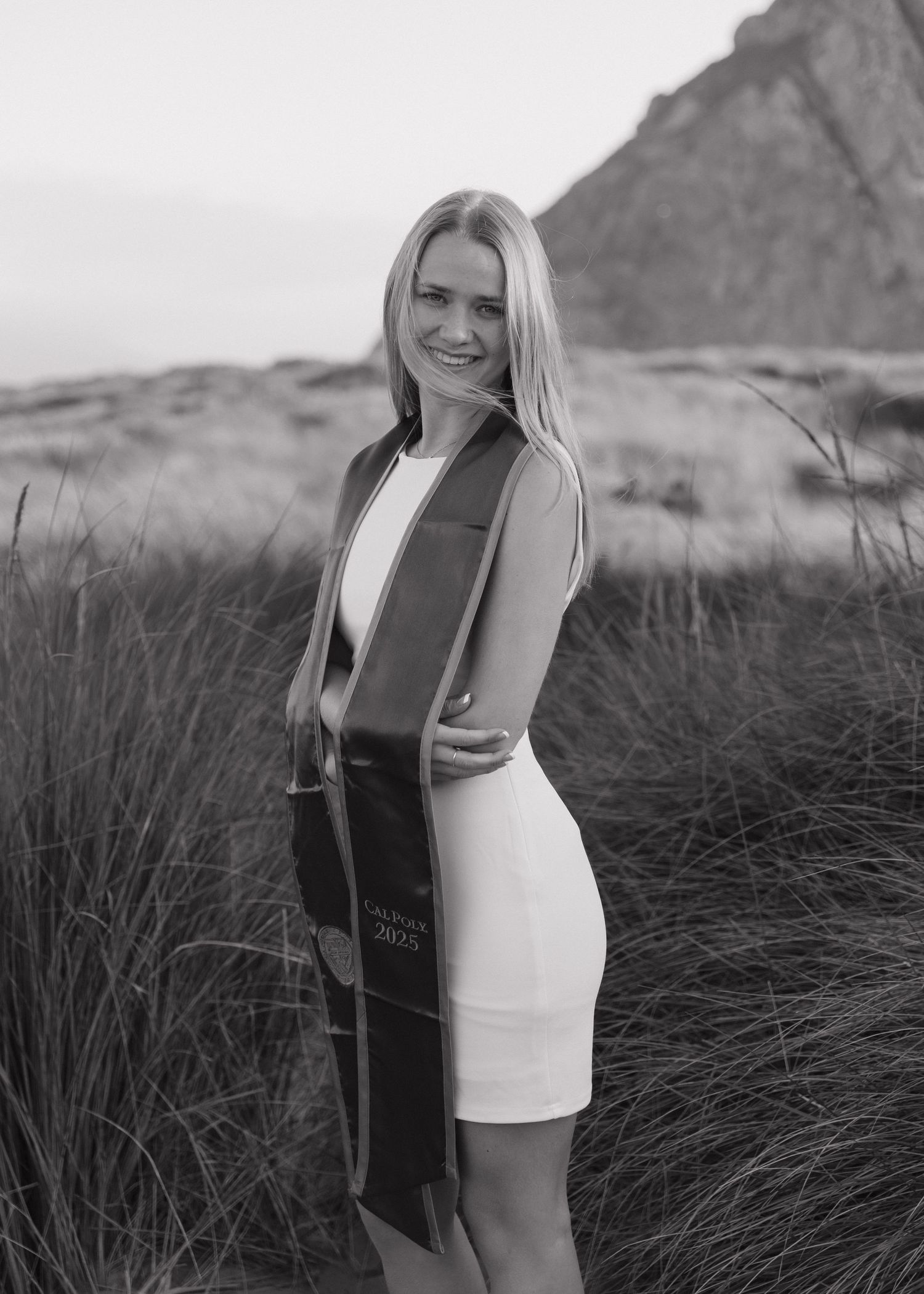 A Cal Poly San Luis Obispo graduate in a white dress poses in tall grass against the Morro Bay Rock In California at sunset.